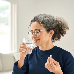 Happy healthy woman of middle age talking pill holding glass of water standing at home. Mature 50 years old lady taking vitamin health care supplement morning daily nutrition treatment concept.