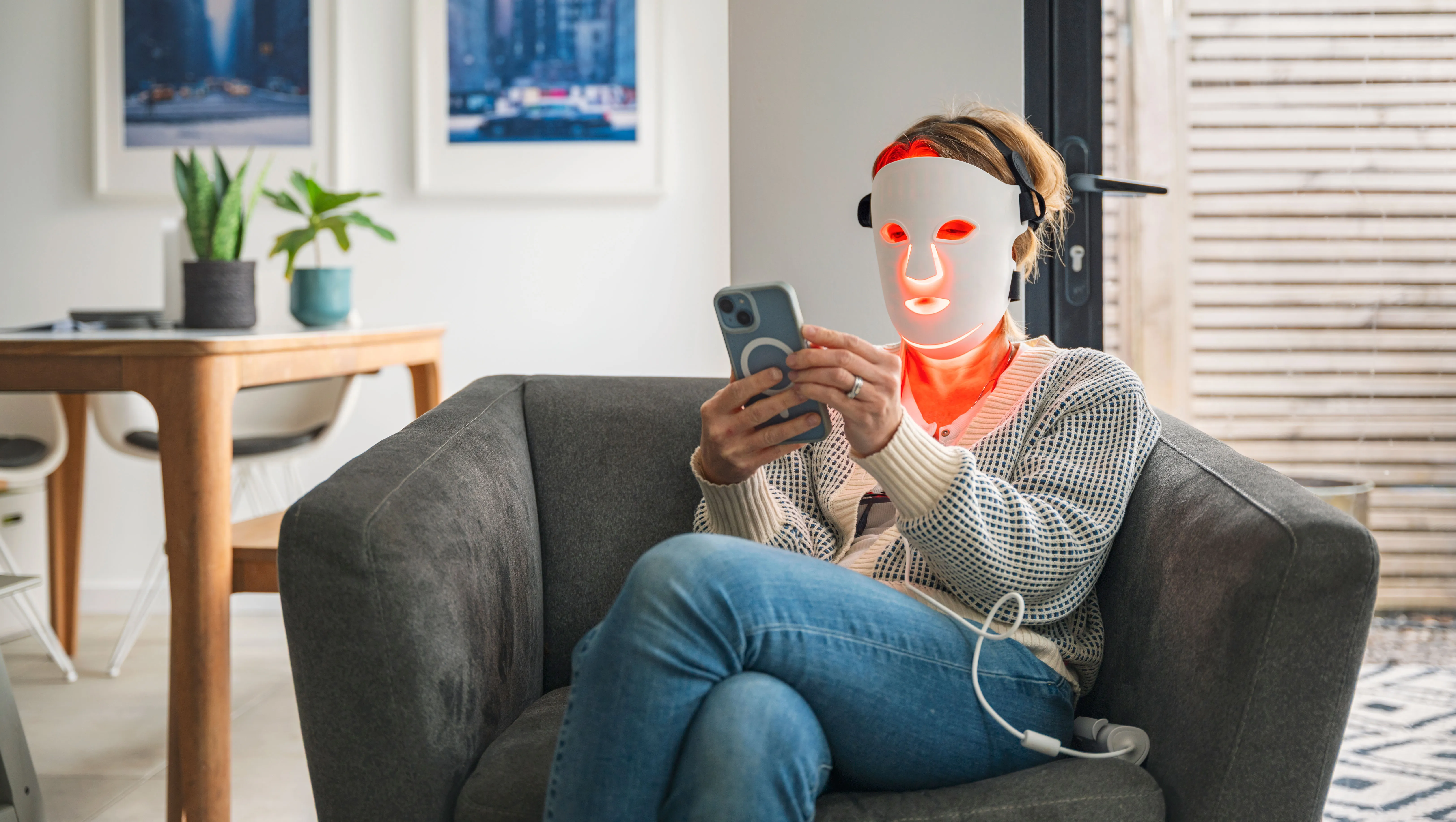 A mature woman uses her red LED face mask (red light therapy) whilst relaxing at home in an armchair. She uses her smartphone - stock photo