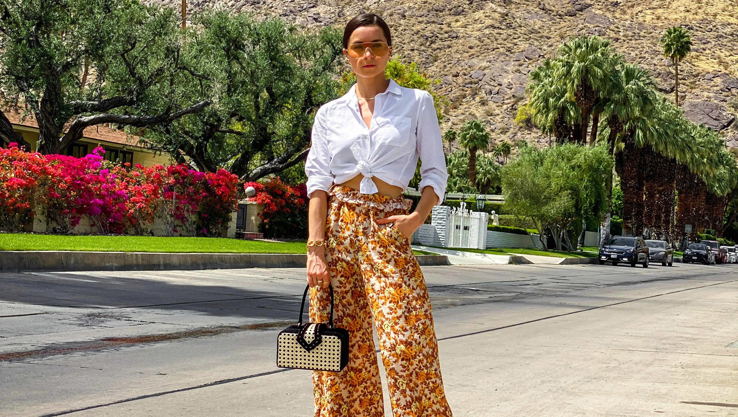 PARIS, FRANCE - APRIL 11: Julia Comil wears floral print flared flowy orange and brown palazzo pants by Jonathan Simkhai, a white linen shirt by Sezane, sustainable mules shoes in biodegradable nappa leather and natural wood by Nicholas Kirkwood, a vintage inspired cane box handbag by Mehry Mu, orange aviator sunglasses by ferragamo, during an online remote street style fashion photo session via Apple iphone / Facetime and the CLOS app as the model is based in Los Angeles - California and the photographer in Paris - France, on April 11, 2021 in Paris, France. (Photo by Edward Berthelot/Getty Images)