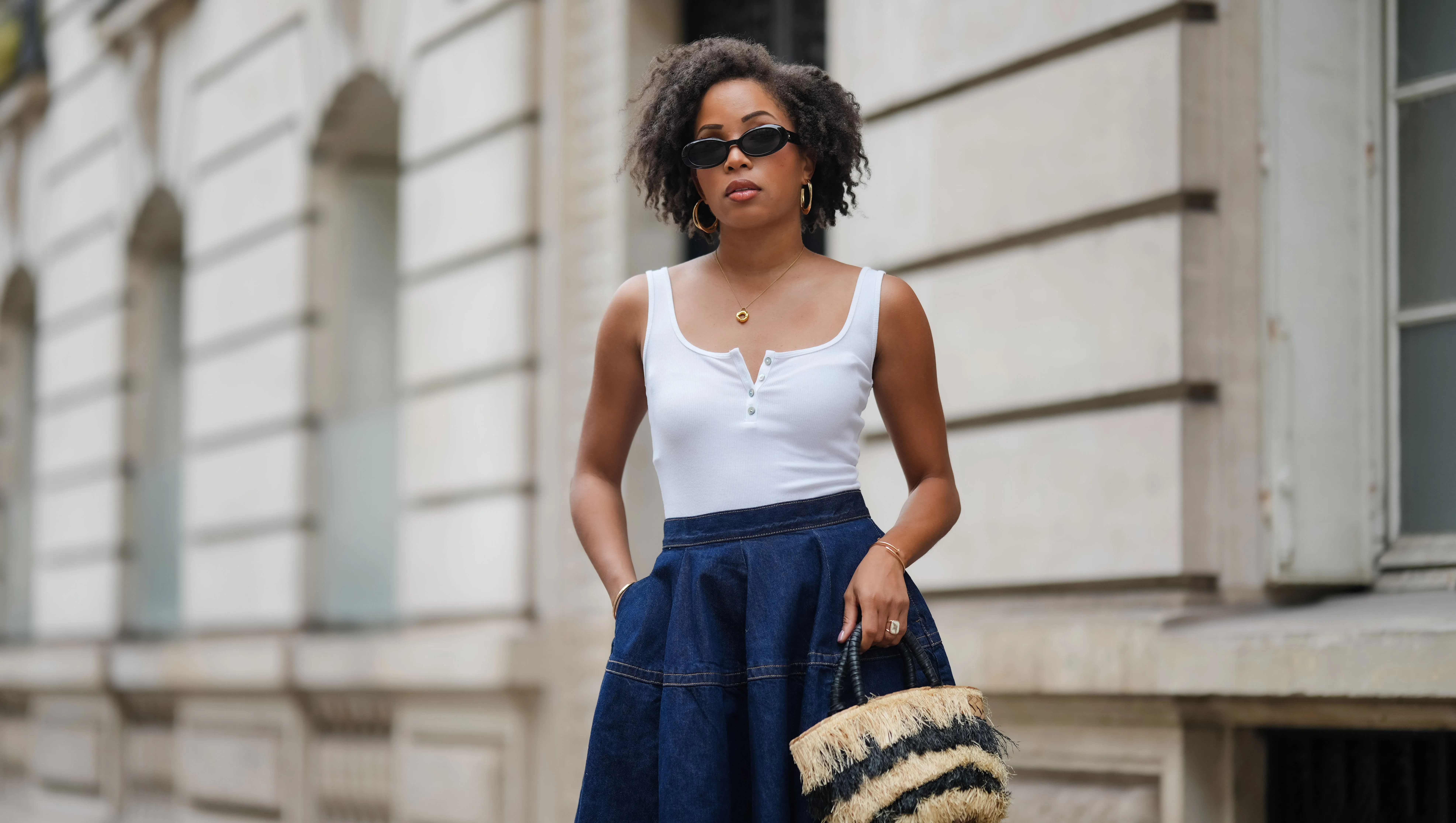 PARIS, FRANCE - MAY 08: Ellie Delphine wears sunglasses , golden earrings, a necklace, a white tank top, a blue midi denim gathered skirt, a beige and black striped fluffy bag, during a street style fashion photo session, on May 08, 2024 in Paris, France. (Photo by Edward Berthelot/Getty Images)