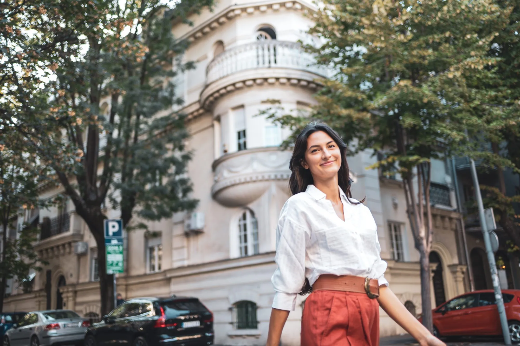A confident woman strolls down a city street lined with lush trees and a historic building, exuding positivity and independence. The scene captures a blend of modern life and architectural charm.