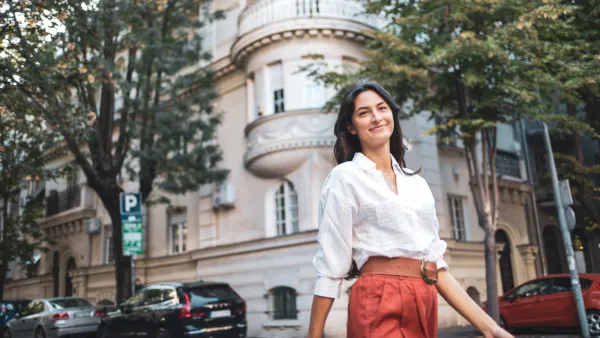 A confident woman strolls down a city street lined with lush trees and a historic building, exuding positivity and independence. The scene captures a blend of modern life and architectural charm.