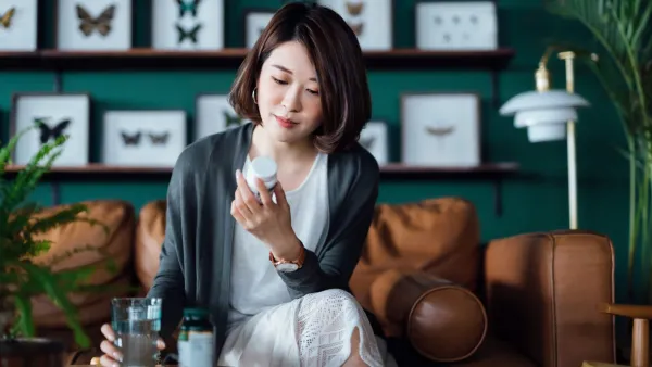 Young Asian woman taking medicines with a glass of water on the coffee table, reading the information on the label of her medication at home. Healthcare concept