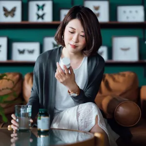 Young Asian woman taking medicines with a glass of water on the coffee table, reading the information on the label of her medication at home. Healthcare concept