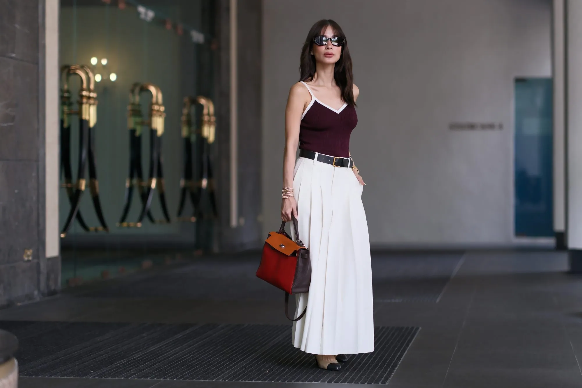 Heart Evangelista wears sunglasses from Dior, a burgundy low-neck tank top with white lines from Edition, a black leather belt, a white maxi long pleated skirt from Edition, golden bracelets, a brown / orange / red leather Kelly Hermes bag, Chanel shoes, during a street style fashion photo session, on November 15, 2023 in Singapore.