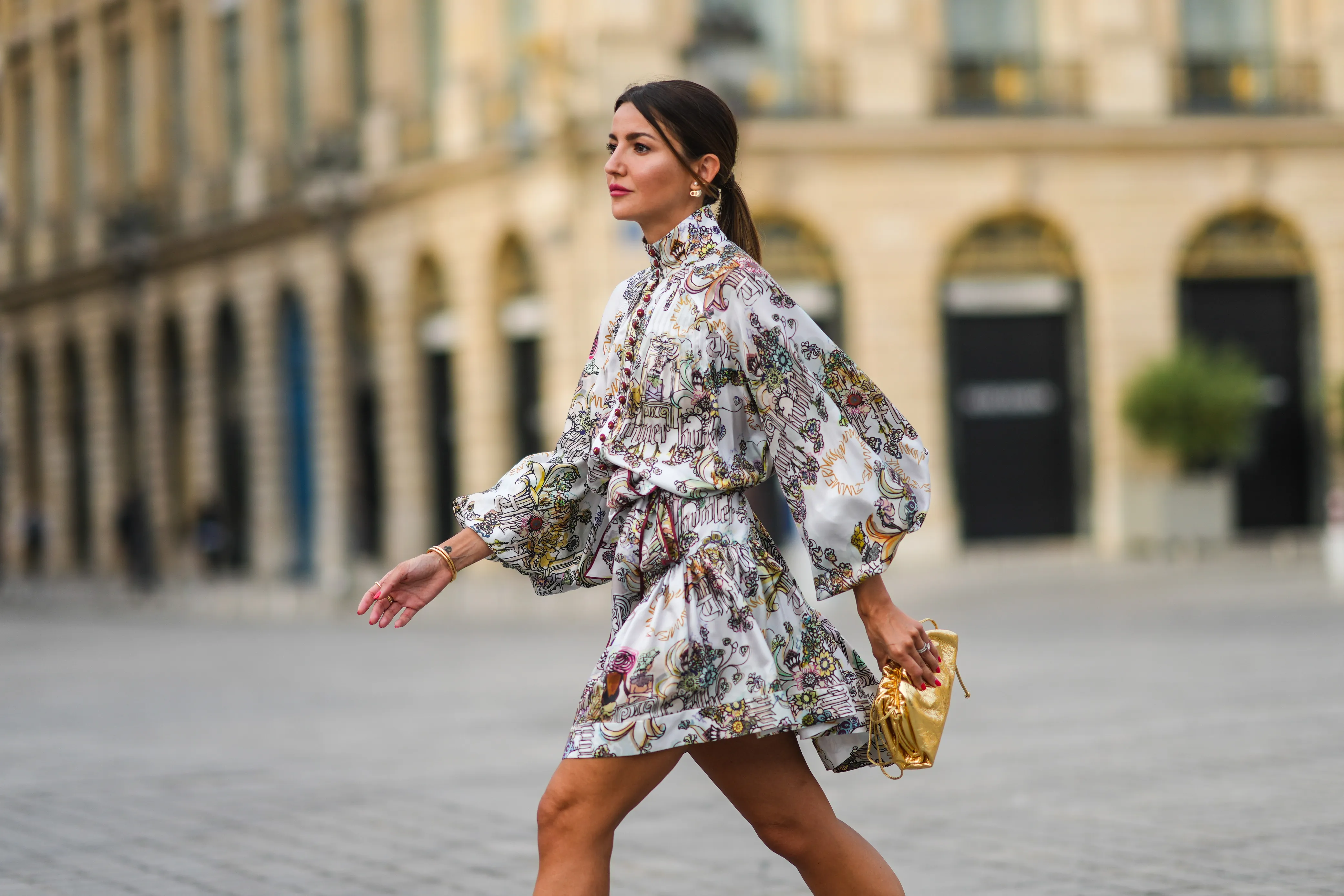 Alexandra Pereira wears gold and pearl earrings, a white with burgundy and pale colors print pattern belted turtleneck dress from Zimmerman with puffy sleeves and burgundy buttons, silver and diamonds rings, gold bracelets from Piaget, a gold shiny leather handbag / clutch from Bottega Veneta, on June 21, 2021 in Paris, France.