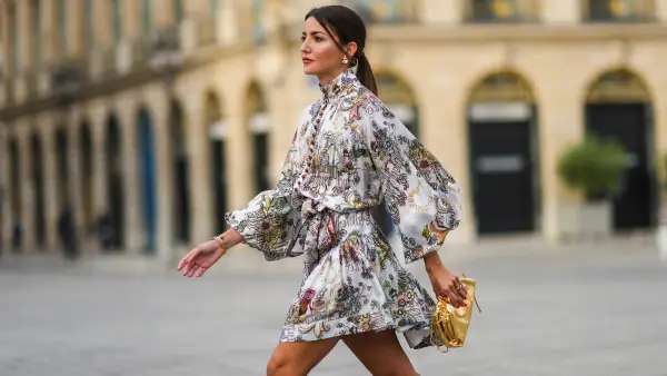 Alexandra Pereira wears gold and pearl earrings, a white with burgundy and pale colors print pattern belted turtleneck dress from Zimmerman with puffy sleeves and burgundy buttons, silver and diamonds rings, gold bracelets from Piaget, a gold shiny leather handbag / clutch from Bottega Veneta, on June 21, 2021 in Paris, France.