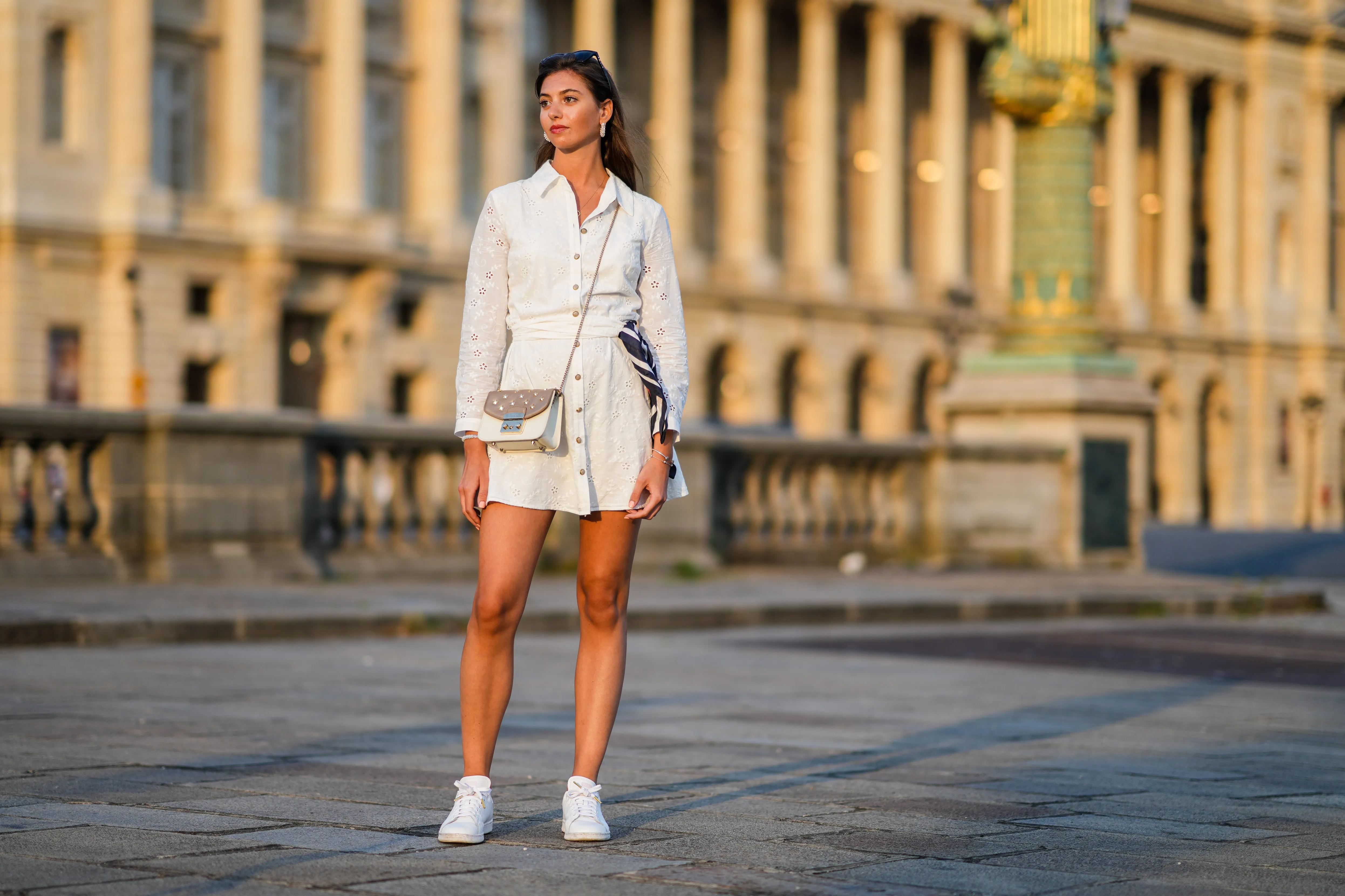 Amanda Derhy wears black sunglasses, pearls pendant earrings, a gold chain pendant necklace, a white cut-out and lace flower pattern buttoned / shirt / belted short dress, a red with navy blue and white striped scarf, silver bracelets, a white moon stones bracelet, a beige matte and brown shiny leather with silver stars nailed / studded crossbody bag from Furla, white with pale pink heart Stan Smith sneakers from Adidas, on August 21, 2021 in Paris, France.