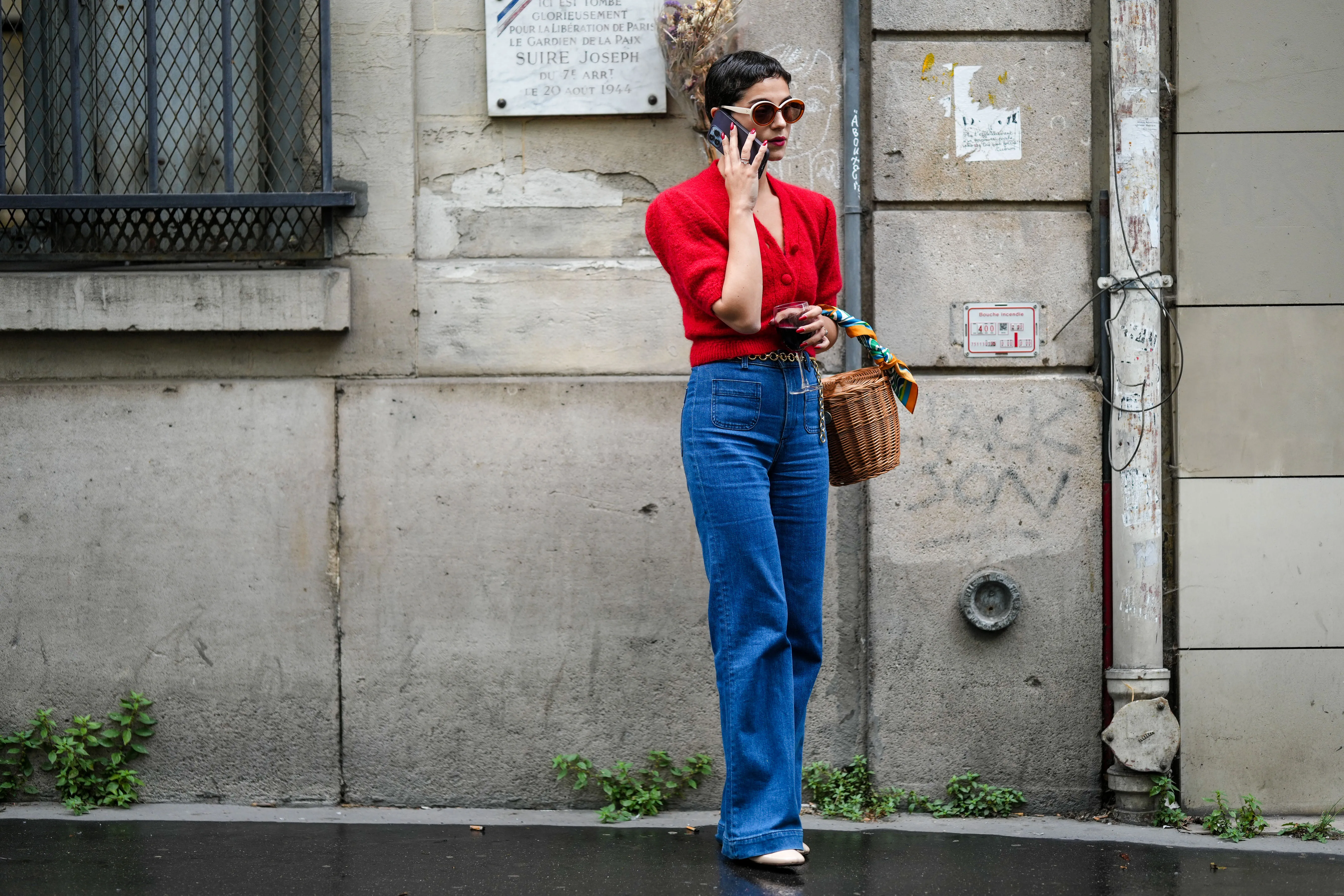 A guest wears circle sunglasses, a red fluffy puffy short sleeves / buttoned cropped cardigan, navy blue denim wide legs pants, a brown wickers basket handbag, a green / blue / orange print pattern silk scarf, rings, white shiny varnished leather heels shoes, a gold chain belt, outside the Hermes show, during Paris Fashion Week - Menswear Spring/Summer 2023, on June 25, 2022 in Paris, France.