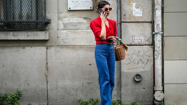 A guest wears circle sunglasses, a red fluffy puffy short sleeves / buttoned cropped cardigan, navy blue denim wide legs pants, a brown wickers basket handbag, a green / blue / orange print pattern silk scarf, rings, white shiny varnished leather heels shoes, a gold chain belt, outside the Hermes show, during Paris Fashion Week - Menswear Spring/Summer 2023, on June 25, 2022 in Paris, France.