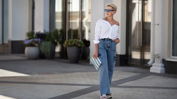 Rejuvenated Trendy mature woman with short hair stands outdoors in city street wears youth clothing white shirt, jeans cargo pants, clutch. Urban style fashion, age and timeless beauty.