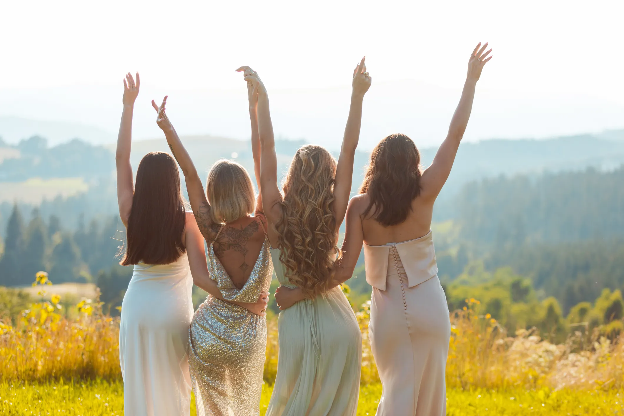 Rear view of female friends in elegant dresses looking at the mountains, embracing and raising arms at sunset. Young women during bachelorette party.