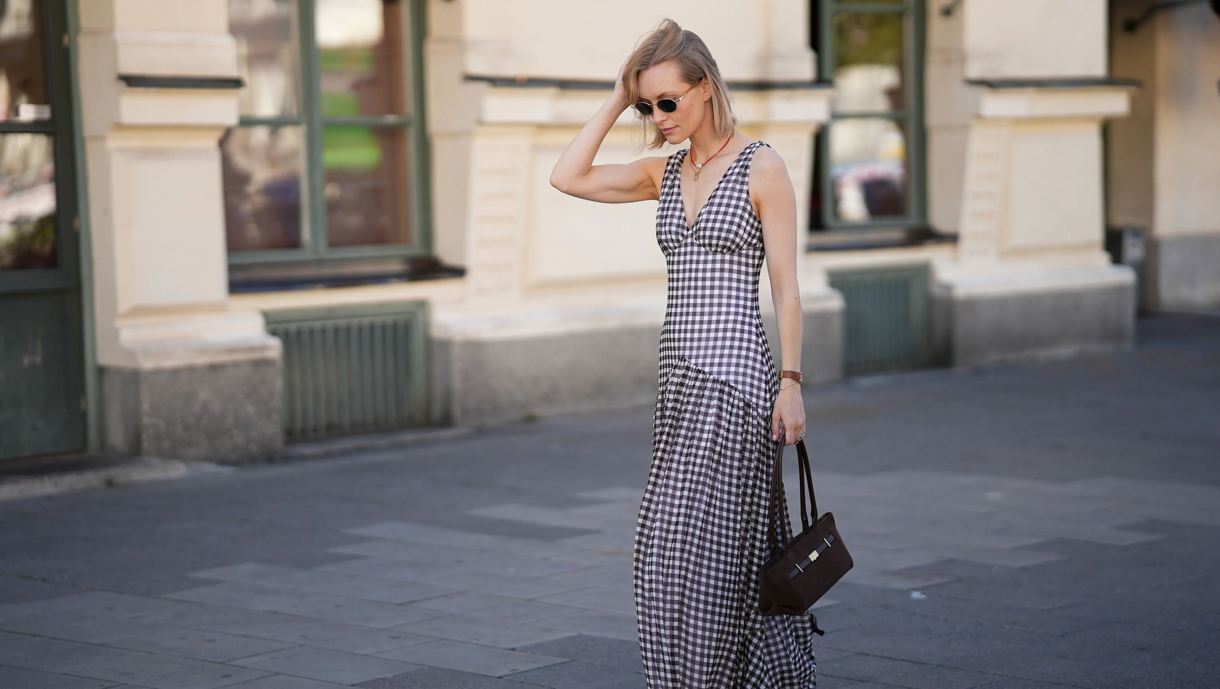 MUNICH, GERMANY - JULY 18: Marlies Pia Pfeifhofer is seen wearing a brown and white Toulouse gingham check maxi dress with a V-neckline, a high front slit, an A-shaped hip cut and a pleated skirt from ADOORE; a dark brown suede The New York shoulderbag with top handles, triangular fold design and golden hardware from DeMellier; round sunglasses with golden temples and dark lenses from Persol; a surigao pearl necklace with red Japanese glass beads and a freshwater baroque pearl from Les Beads Studio; a golden medallion necklace with faceted diamond bezel and hand-painted 'Porte Bonheur' enamel from Marlo Laz; small golden hoop earrings from Monica Vinader; a vintage square Tank wristwatch with light brown leather strap from Cartier; a diamond ring; her blonde hair is styled in a straight bob with a center part on July 18, 2025 in Munich, Germany. (Photo by Moritz Scholz/Getty Images)