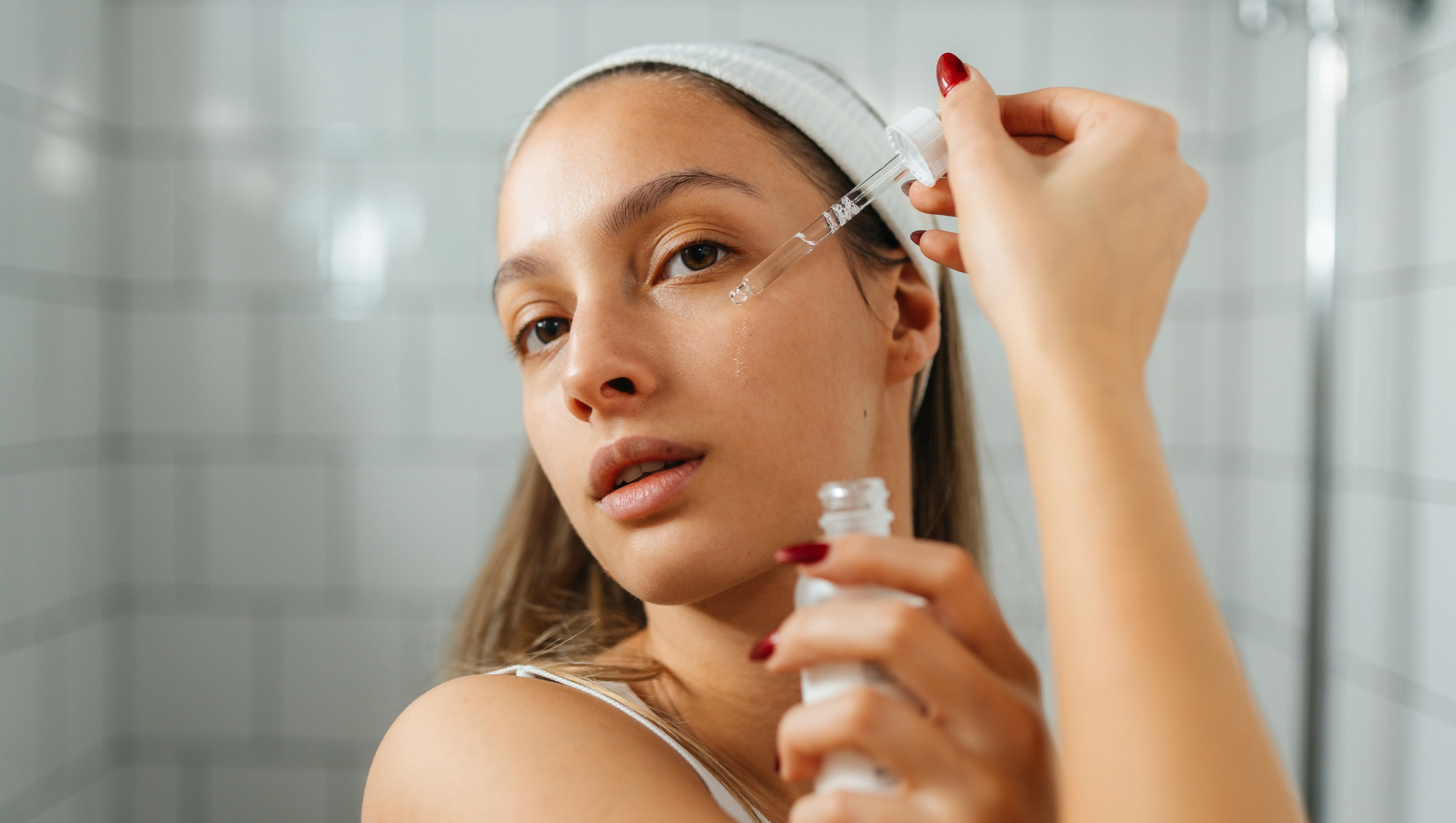 Young woman standing in a bright, modern bathroom, applying facial serum to her skin. She holds a small dropper bottle in one hand and gently applies the serum to her face.