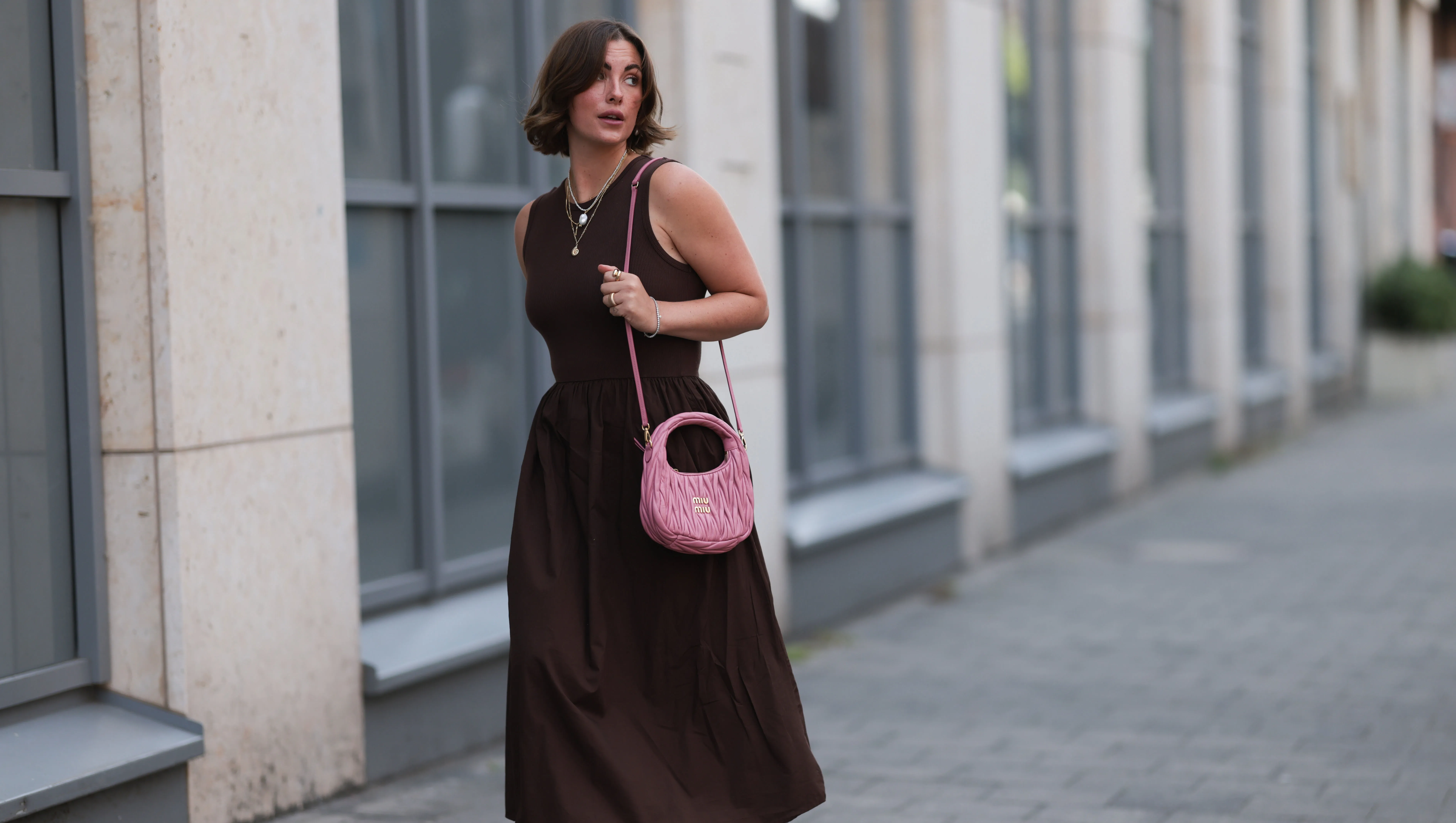 HAMBURG, GERMANY - JULY 18: Franzi Koenig is seen wearing several stud earrings, including gold seashell-shaped studs from fafe collection; layered gold necklaces with a seashell pendant, an F-letter pendant, and a white beaded necklace with a round organic-shaped pendant, also from fafe collection; a sleeveless dark brown midi dress with a ribbed top, high round neckline, and a gathered A-line skirt; a quilted pink leather “Wander” Hobo bag with gold logo hardware and a long thin shoulder strap from Miu Miu; several chunky gold rings, paired with delicate bracelets, both from fafe collection; and dark brown slip-on sandals with kitten heels on July 18, 2025 in Hamburg, Germany. (Photo by Jeremy Moeller/Getty Images)