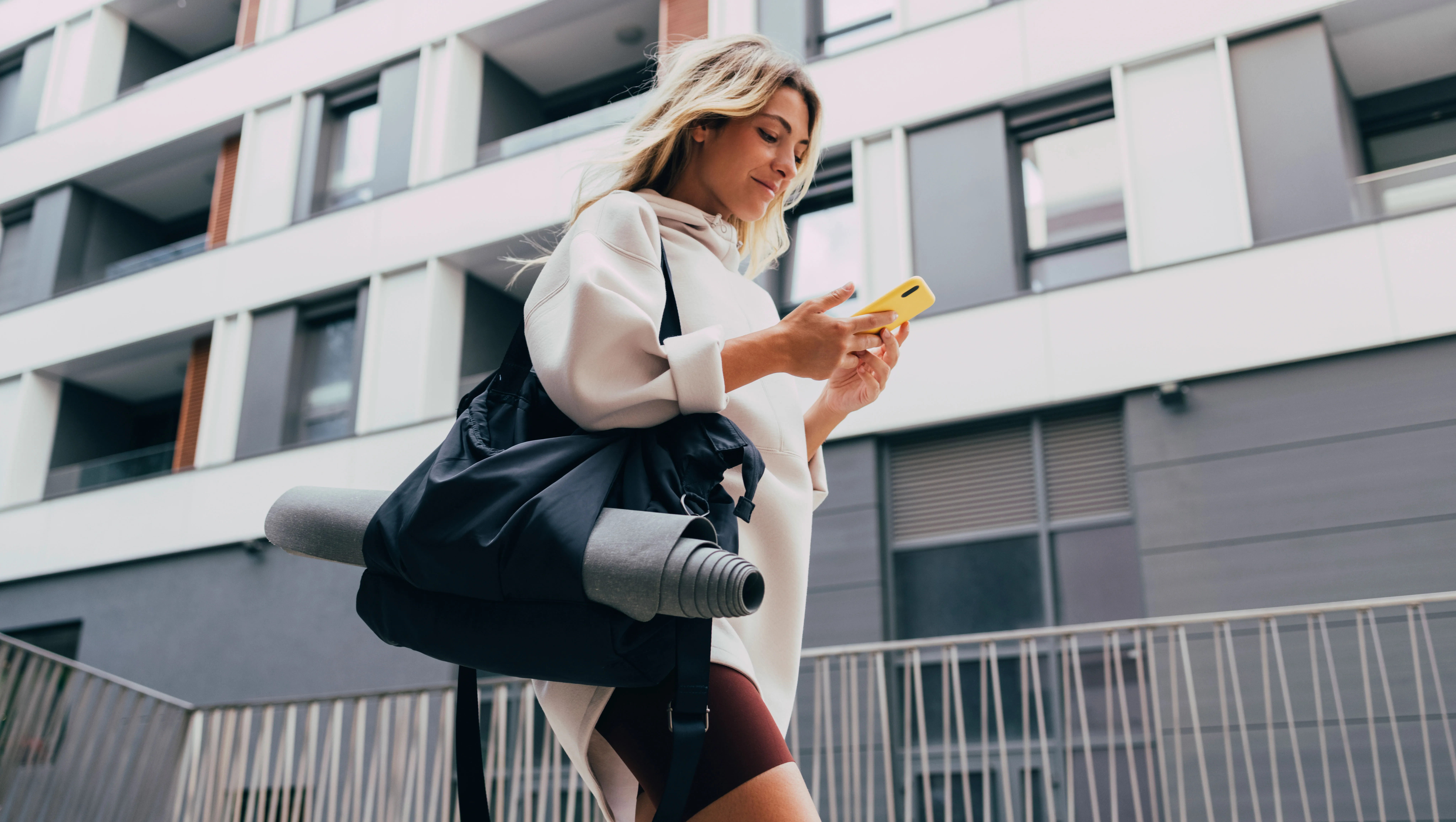 Cheerful smiling woman in sportswear typing text message on her smartphone while walking in the city