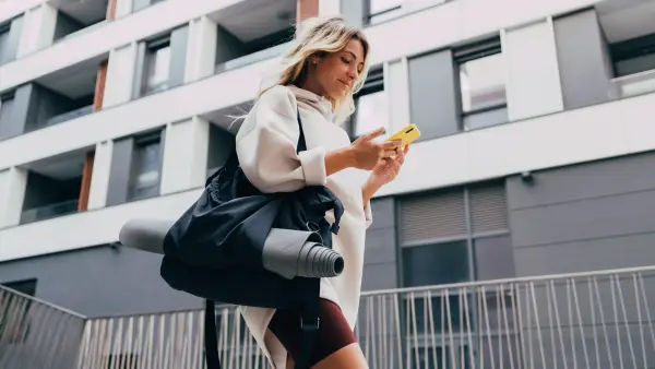 Cheerful smiling woman in sportswear typing text message on her smartphone while walking in the city