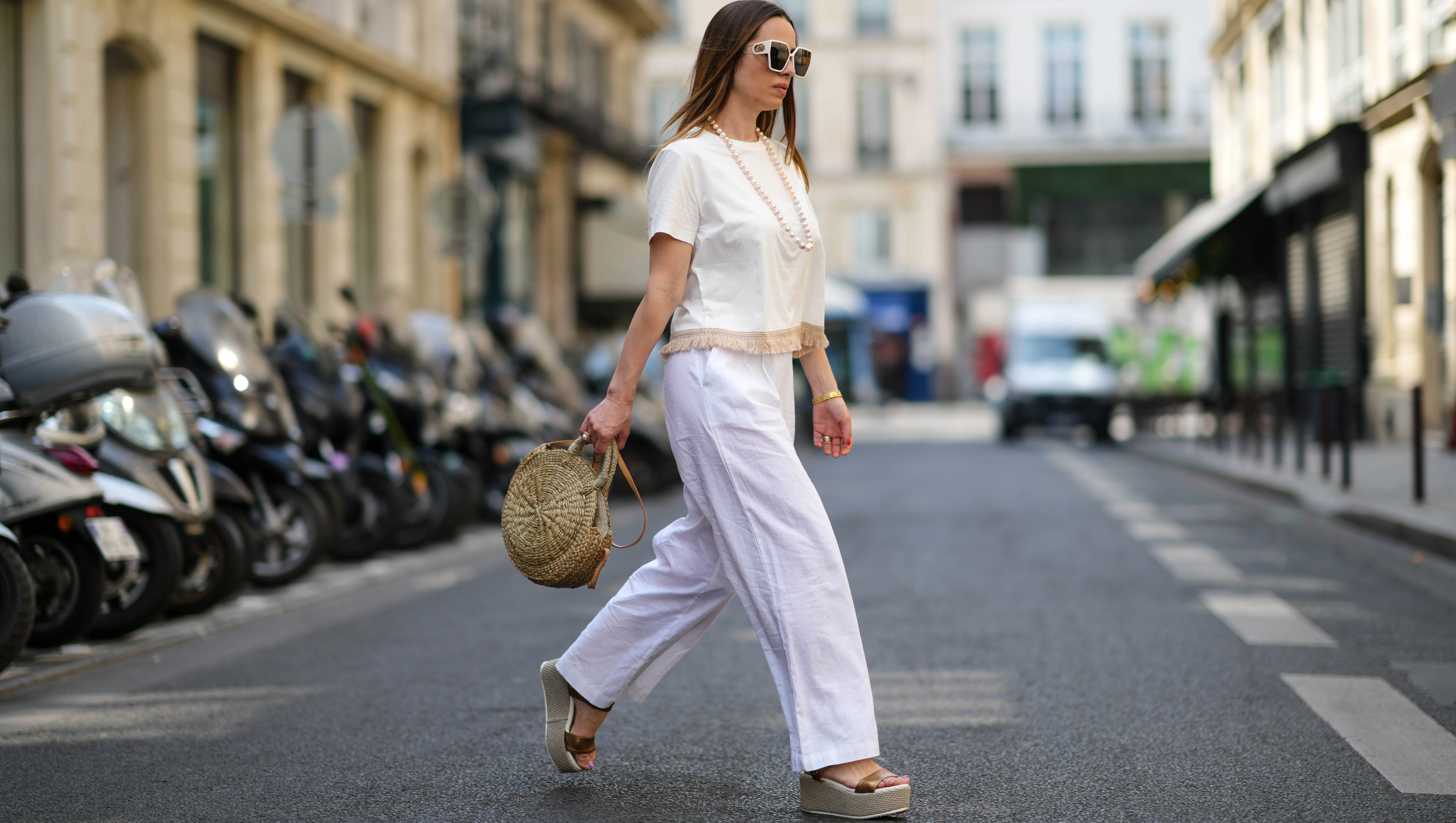 PARIS, FRANCE - MAY 10: Maria Rosaria Rizzo wears white sunglasses, a white pearls long necklace, a white latte t-shirt with fringed borders, a beige wickers handbag from Guerlain, a gold large bracelet, gold rings, high waist white wide legs linen pants, gold shiny leather strappy with wicker wedge heels sandals, during a street style fashion photo session, on May 10, 2022 in Paris, France. (Photo by Edward Berthelot/Getty Images)