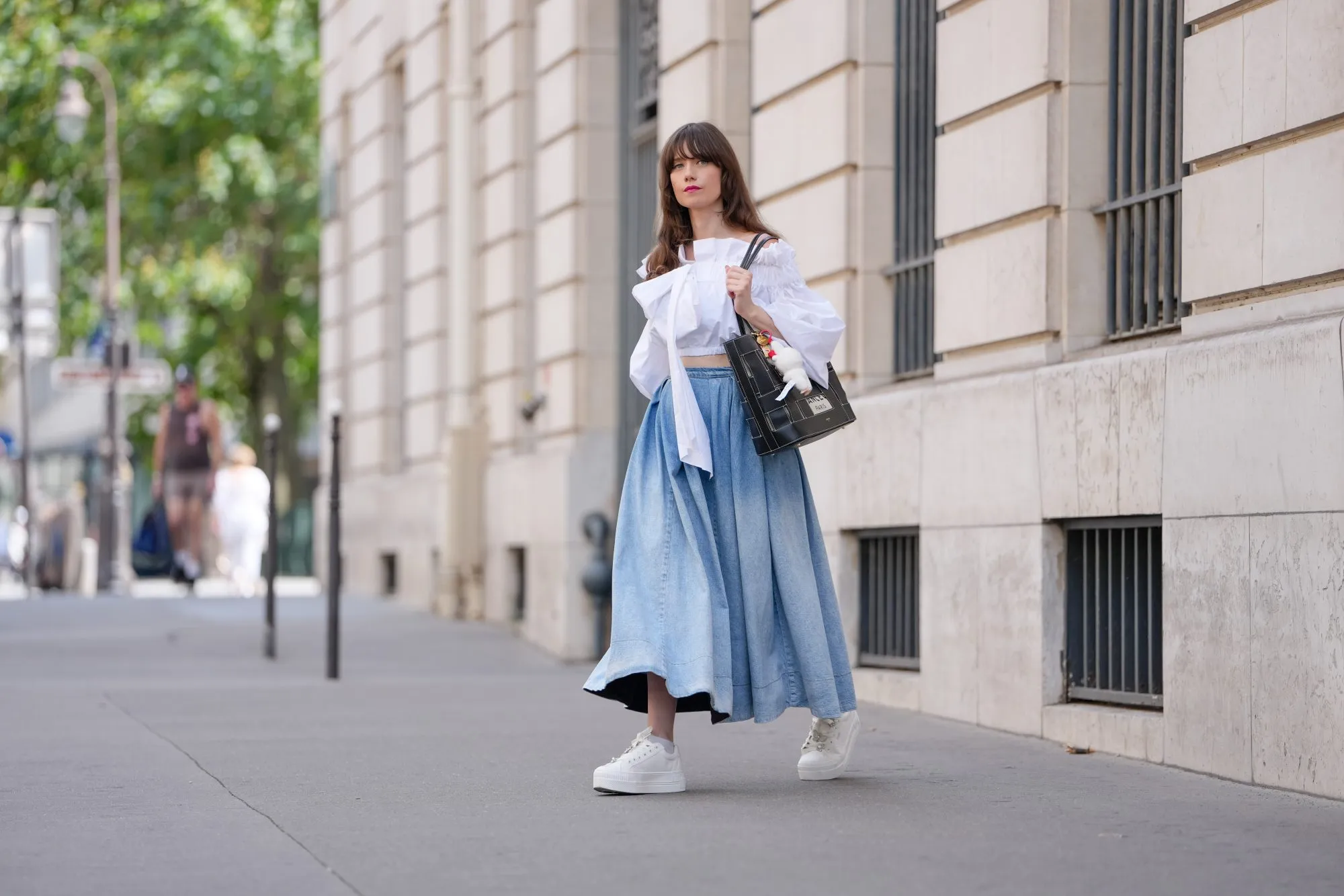 PARIS, FRANCE - JULY 18: Marion Félicité wears a white cropped top with oversized puffed sleeves, gathered seams and an asymmetrical bow detail on the chest by Patou, paired with a high-waisted, voluminous light blue denim maxi skirt with washed effect. A black and beige woven leather tote bag by Lancel is carried by hand, customized with a "Surprise Shake" Labubu Coca-Cola plush charm and colorful enamel keyrings. White Buffalo platform sneakers with lace-up closure are adorned with decorative silver and pearl charms including a butterfly and floral motif. Multicolored beaded bracelets are worn on the wrist. Fingernails are painted in a glossy gradient pink with green. Hair is styled in long, loose waves with front bangs and a side part. during a street style fashion photo session, on July 18, 2025 in Paris, France. (Photo by Edward Berthelot/Getty Images)