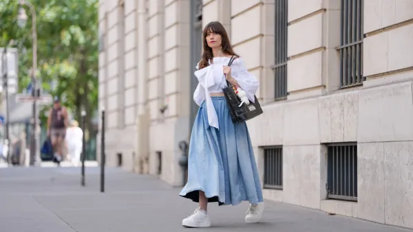 PARIS, FRANCE - JULY 18: Marion Félicité wears a white cropped top with oversized puffed sleeves, gathered seams and an asymmetrical bow detail on the chest by Patou, paired with a high-waisted, voluminous light blue denim maxi skirt with washed effect. A black and beige woven leather tote bag by Lancel is carried by hand, customized with a "Surprise Shake" Labubu Coca-Cola plush charm and colorful enamel keyrings. White Buffalo platform sneakers with lace-up closure are adorned with decorative silver and pearl charms including a butterfly and floral motif. Multicolored beaded bracelets are worn on the wrist. Fingernails are painted in a glossy gradient pink with green. Hair is styled in long, loose waves with front bangs and a side part. during a street style fashion photo session, on July 18, 2025 in Paris, France. (Photo by Edward Berthelot/Getty Images)