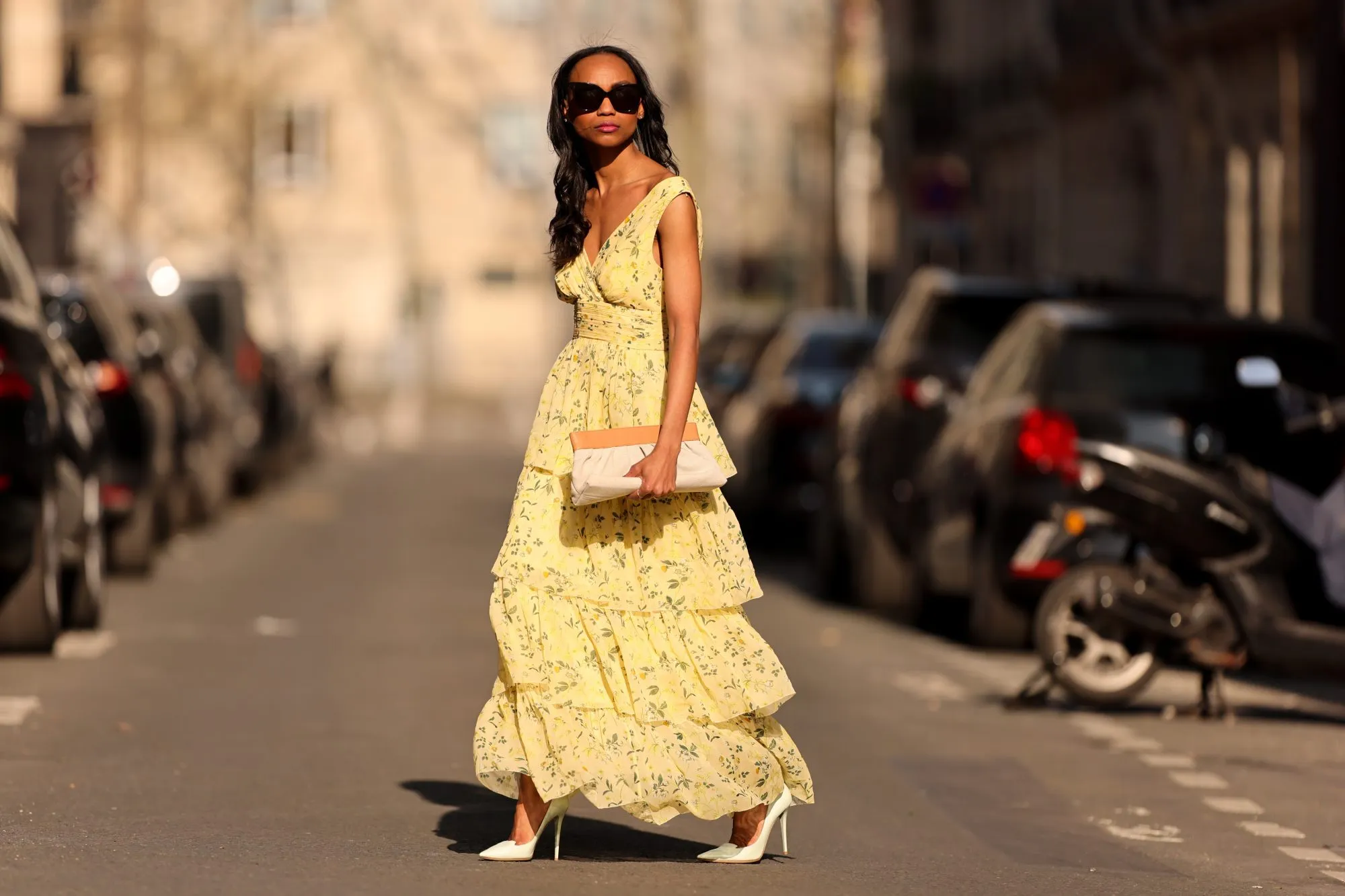 PARIS, FRANCE - MARCH 26: Emilie Joseph @in_fashionwetrust wears black sunglasses, a pale yellow with green and yellow flower print pattern V-neck / tank-top / backless / belted / ruffled maxi long dress, a beige fabric ruffled clutch, silver rings, pale yellow matte leather pointed pumps heels shoes, during a street style fashion photo session, on March 26, 2022 in Paris, France. (Photo by Edward Berthelot/Getty Images)