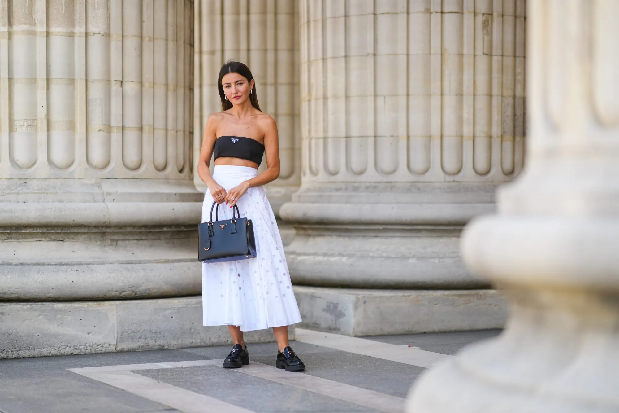 PARIS, FRANCE - JUNE 21: Alexandra Pereira wears silver and gold earrings, a black Prada shoulder-off crop-top, a white high accordion / gathered / pleated midi skirt with round holes from Prada, a black grained leather Galleria from Prada handbag, silver and diamonds rings, black shiny leather loafer shoes from Prada, on June 21, 2021 in Paris, France. (Photo by Edward Berthelot/Getty Images)