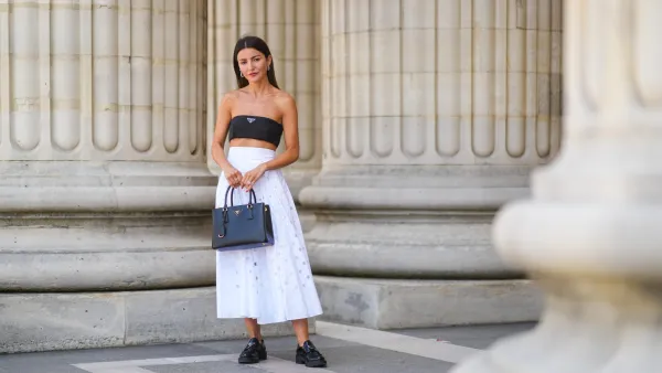 PARIS, FRANCE - JUNE 21: Alexandra Pereira wears silver and gold earrings, a black Prada shoulder-off crop-top, a white high accordion / gathered / pleated midi skirt with round holes from Prada, a black grained leather Galleria from Prada handbag, silver and diamonds rings, black shiny leather loafer shoes from Prada, on June 21, 2021 in Paris, France. (Photo by Edward Berthelot/Getty Images)
