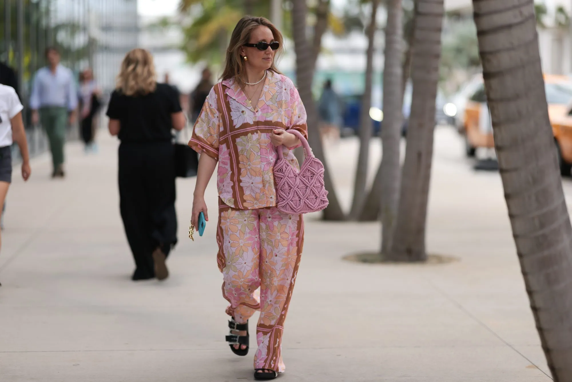 MIAMI, FLORIDA - DECEMBER 08: Anna Sophie seen wearing Le Specs brown oval sunglasses, gold earrings and necklace, pearl necklace, pink colorful floral print pattern shirt / blouse, matching pink colorful floral print pattern long pants, pink crochet handbag and black leather sandals, on December 08, 2023 in Miami, Florida. (Photo by Jeremy Moeller/Getty Images)