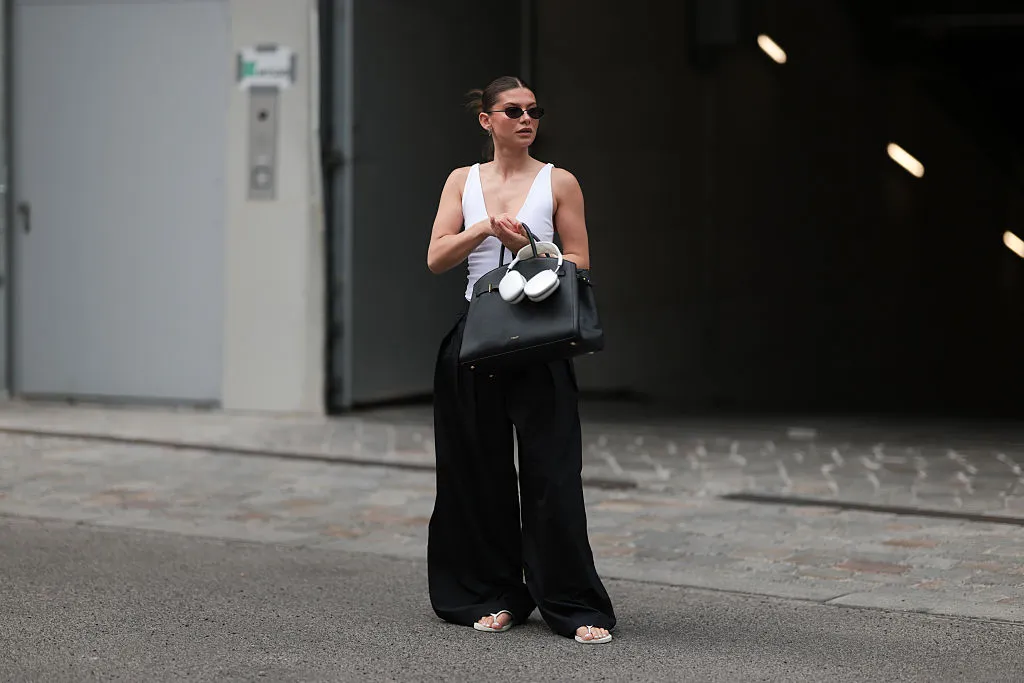 BERLIN, GERMANY - JUNE 04: Sophia Geiss is seen wearing silver hoop earrings in two different sizes; oval, rimless black sunglasses from Vehla Eyewear; a silver nose ring; a white sleeveless bodysuit with a deep neckline from Zara; pleated wide-leg black suit pants from COS; a large rectangular black leather handbag from DeMellier; a gold bangle paired with a chunky beige bangle; and white flip-flops from Havaianas, while holding silver Apple AirPods Max headphones on June 04, 2025 in Berlin, Germany. (Photo by Jeremy Moeller/Getty Images)
