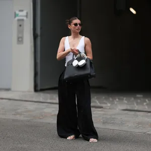 BERLIN, GERMANY - JUNE 04: Sophia Geiss is seen wearing silver hoop earrings in two different sizes; oval, rimless black sunglasses from Vehla Eyewear; a silver nose ring; a white sleeveless bodysuit with a deep neckline from Zara; pleated wide-leg black suit pants from COS; a large rectangular black leather handbag from DeMellier; a gold bangle paired with a chunky beige bangle; and white flip-flops from Havaianas, while holding silver Apple AirPods Max headphones on June 04, 2025 in Berlin, Germany. (Photo by Jeremy Moeller/Getty Images)
