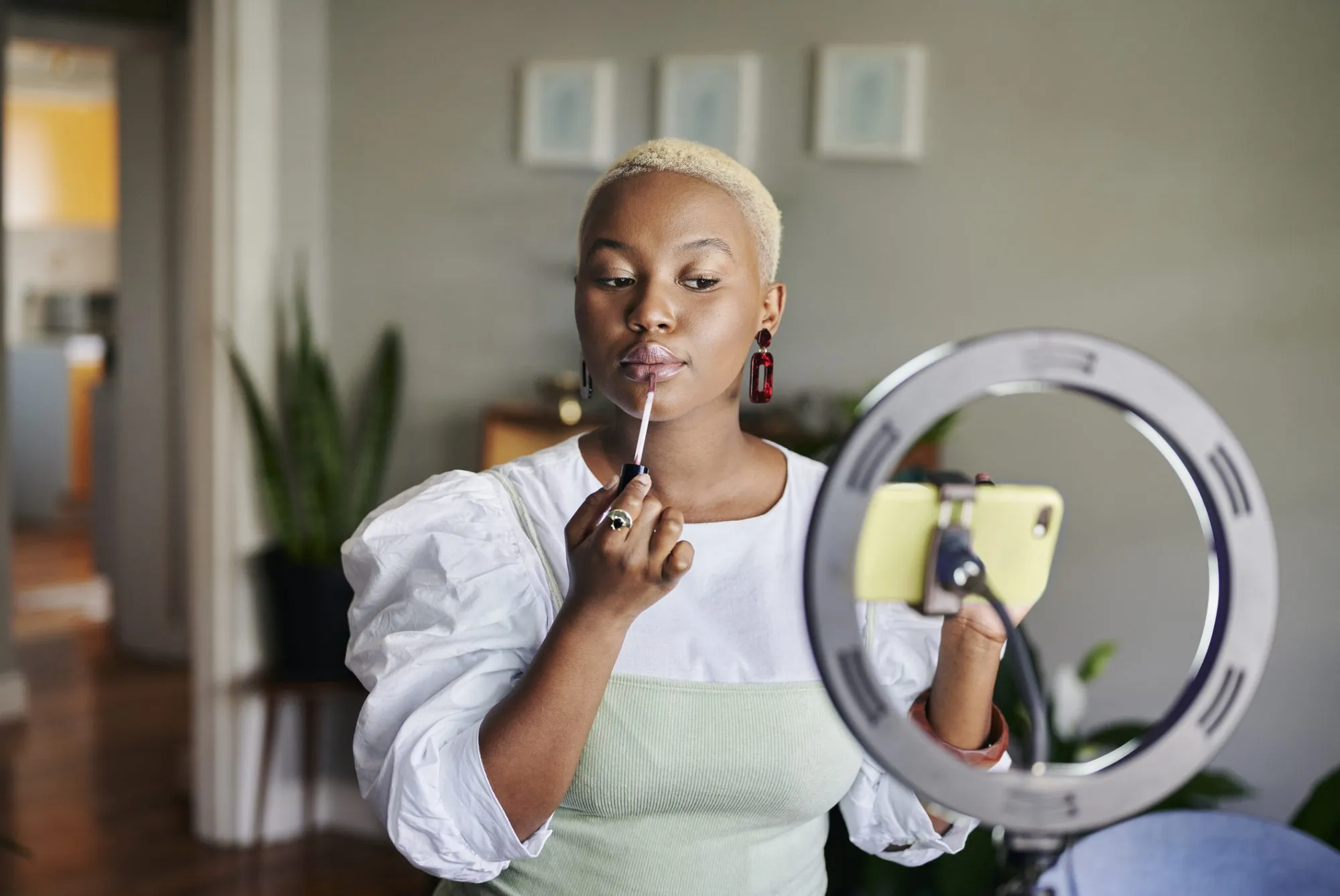 Young African female influencer applying lip gloss while doing a vlogcast from home using a smart phone and ring light. (Photo by mapodile/Getty Images)