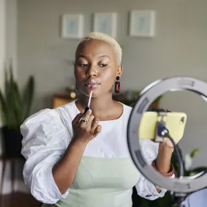 Young African female influencer applying lip gloss while doing a vlogcast from home using a smart phone and ring light. (Photo by mapodile/Getty Images)