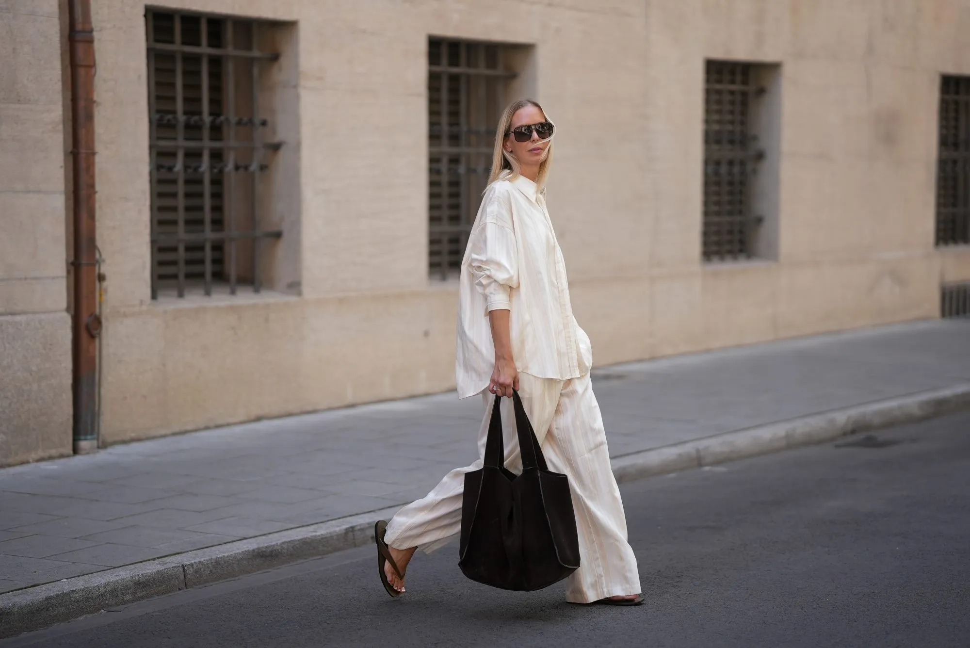 MUNICH, GERMANY - JUNE 18: Mirja Klein is seen wearing a matching set consisting of a white striped oversized dobby-weave shirt with collar and buttons down in the front from H&M and a white striped straight pull-on trousers with loose fit and straight legs from H&M; a black suede tote bag with slouchy design and open top from COS; black oversized aviator sunglasses from Tom Ford; a pair of khaki green suede flip flops from A.EMERY on June 18, 2025 in Munich, Germany. (Photo by Moritz Scholz/Getty Images)