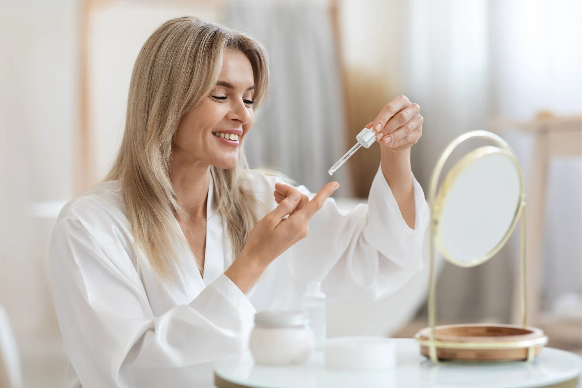Happy beautiful middle aged woman in bathrobe sitting in front of mirror, holding bottle with facial oil or serum, lady applying face care product cosmetics after morning shower, copy space. (Photo by Prostock-Studio/Getty Images)