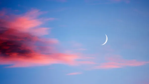 New moon at sunset with light clouds in various shades of red and magenta, darker to the left and a blue sky behind.