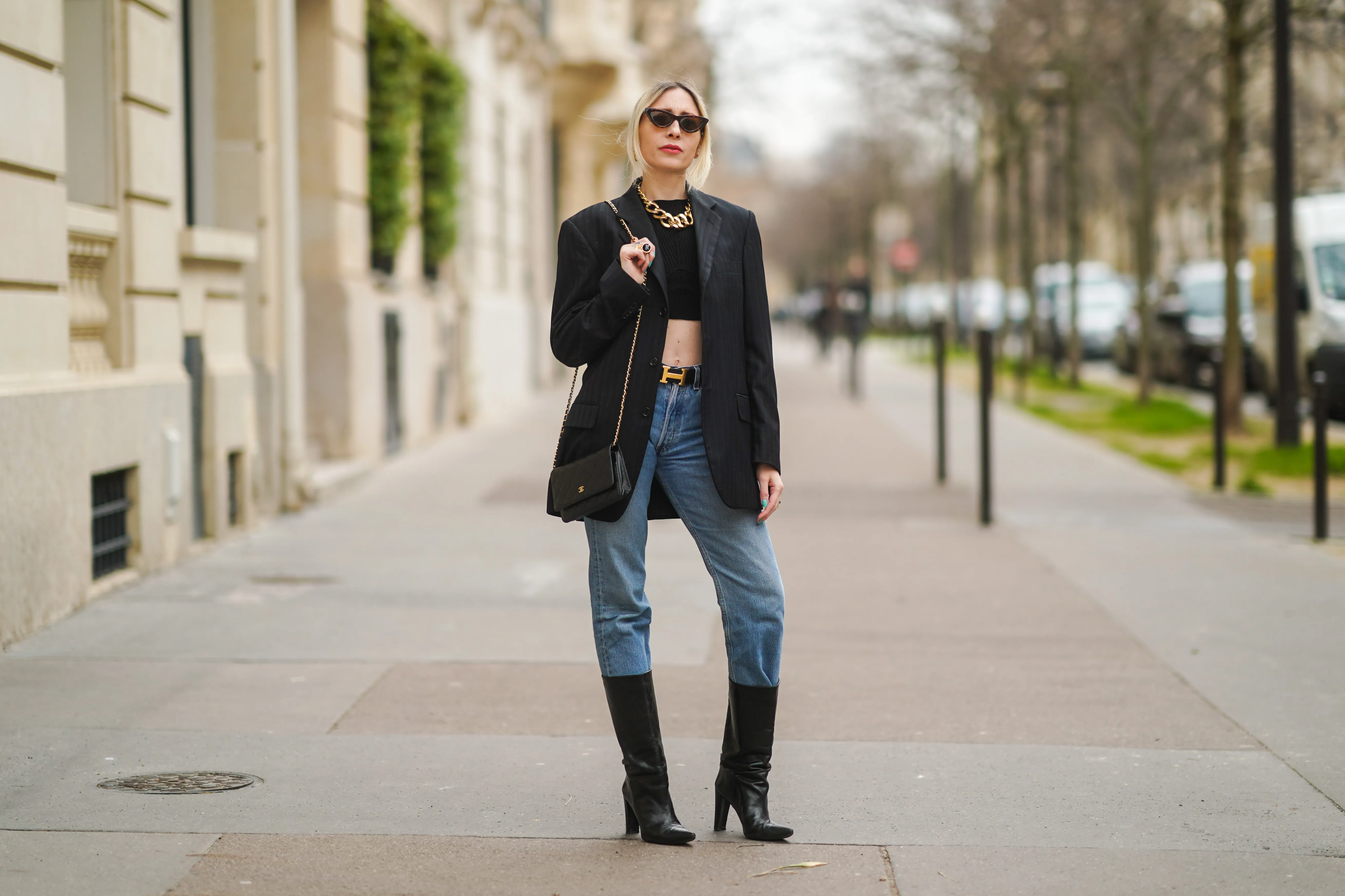 PARIS, FRANCE - FEBRUARY 22: Emy Venturini wears sunglasses, golden earrings, a golden large chain necklace, a black cropped top from Elisabetta Franchi, a black oversized blazer jacket, a Hermes belt, a black leather Chanel bag, blue denim jeans 501 Levi's, black high heeled pointy leather high boots, on February 22, 2021 in Paris, France.