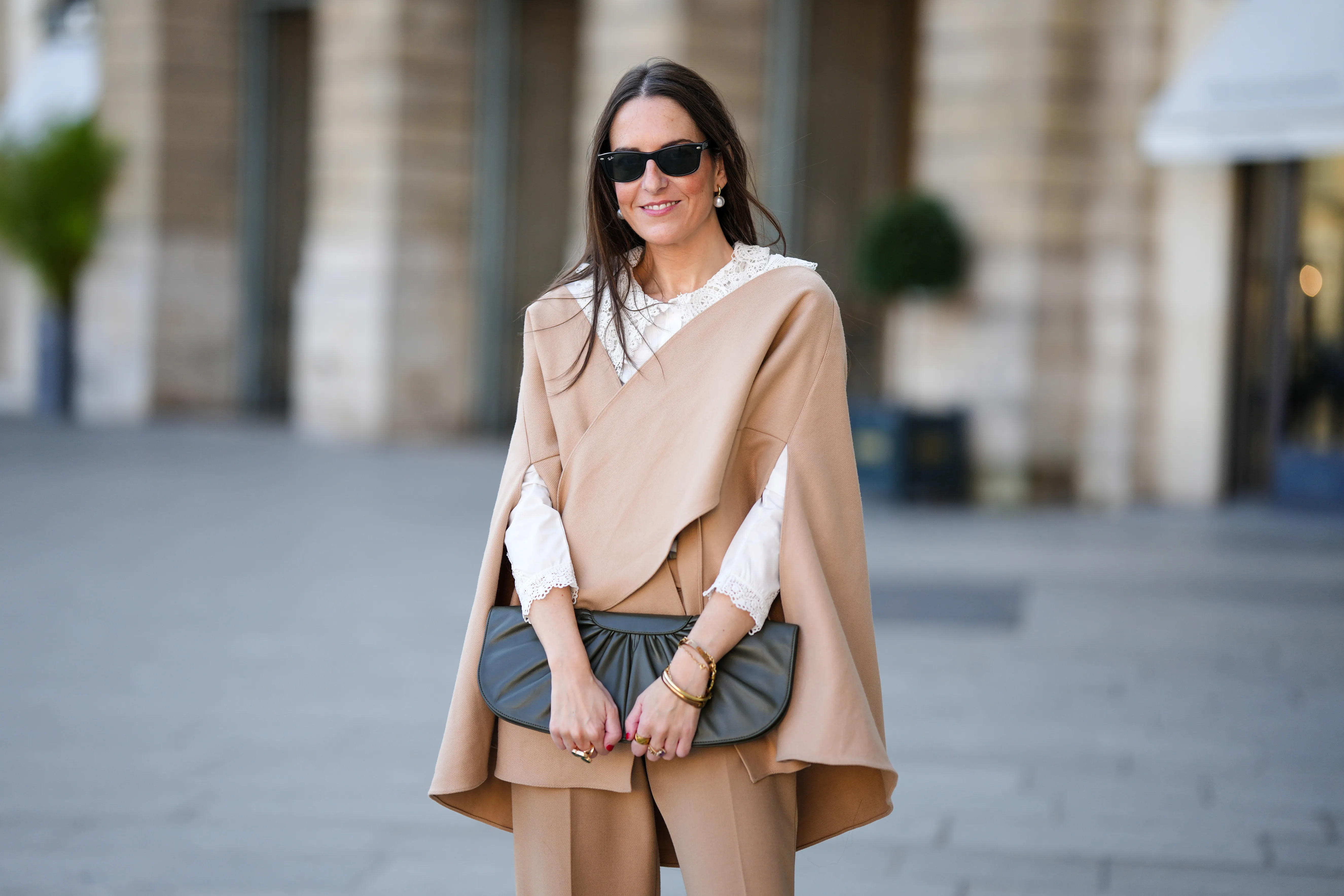 Alba Garavito Torre wears black sunglasses from Ray Ban, white large pearls earrings, a white shirt with cut-out lace pattern large collar, a beige felt / wool oversized cloak, matching beige wool / felt palazzo pants, a black shiny leather ruffled handbag, a gold Love bracelet from Cartier, gold bracelet, gold rings, during a street style fashion photo session, on October 28, 2021 in Paris, France.