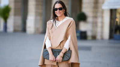 Alba Garavito Torre wears black sunglasses from Ray Ban, white large pearls earrings, a white shirt with cut-out lace pattern large collar, a beige felt / wool oversized cloak, matching beige wool / felt palazzo pants, a black shiny leather ruffled handbag, a gold Love bracelet from Cartier, gold bracelet, gold rings, during a street style fashion photo session, on October 28, 2021 in Paris, France.