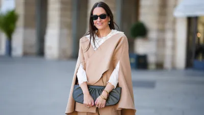 Alba Garavito Torre wears black sunglasses from Ray Ban, white large pearls earrings, a white shirt with cut-out lace pattern large collar, a beige felt / wool oversized cloak, matching beige wool / felt palazzo pants, a black shiny leather ruffled handbag, a gold Love bracelet from Cartier, gold bracelet, gold rings, during a street style fashion photo session, on October 28, 2021 in Paris, France.