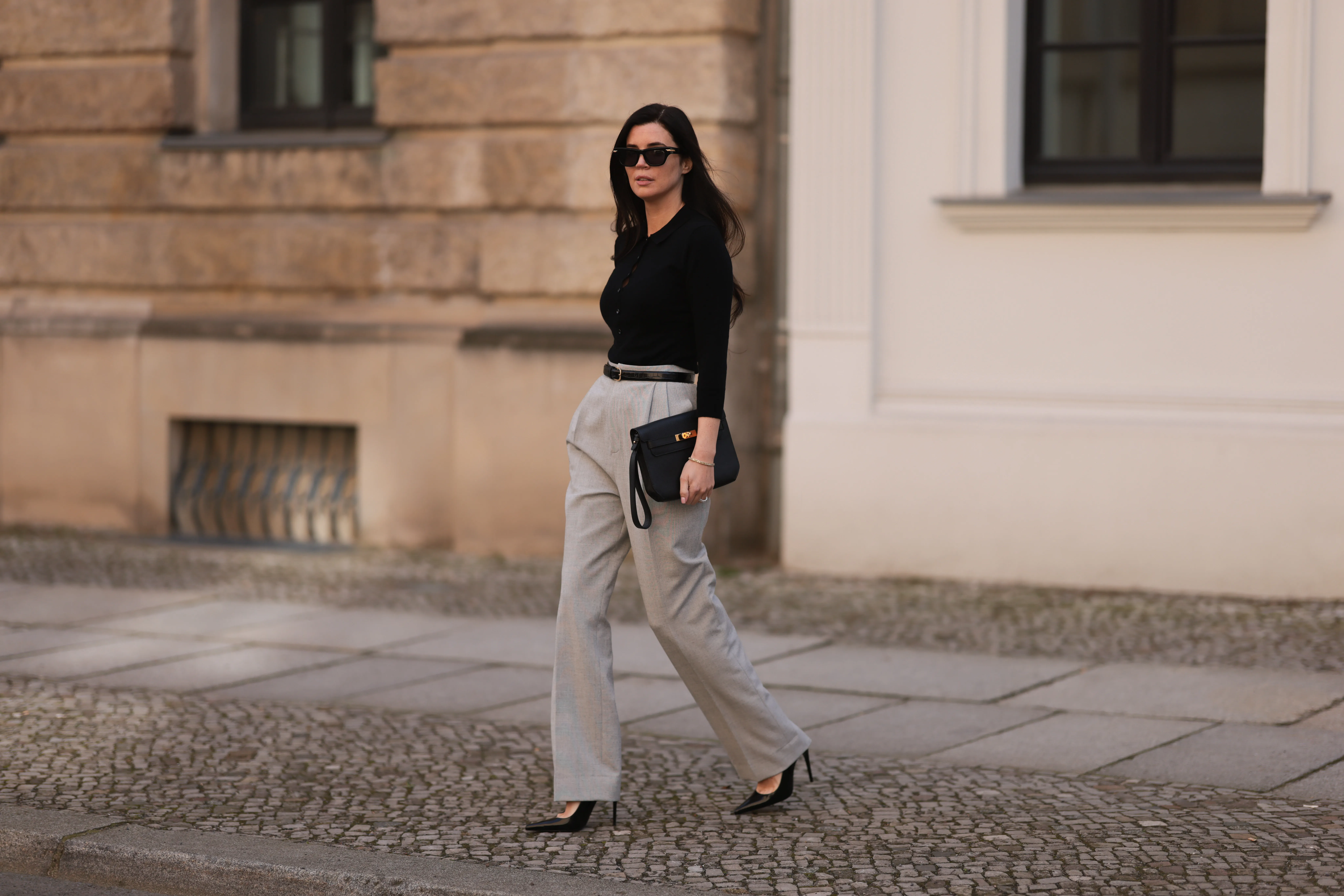 Leo Eberlin seen wearing Bottega Veneta black sunglasses, Leo Mathild jewelry / diamond bracelet and rings, Allude x Leo Mathild cashmere wool knit buttoned body, Toteme black leather belt, Velaa light grey elegant suit pants, Herm&egrave;s black leather clutch bag and Saint Laurent black varnished leather heels / pumps, on February 25, 2024 in Berlin, Germany.