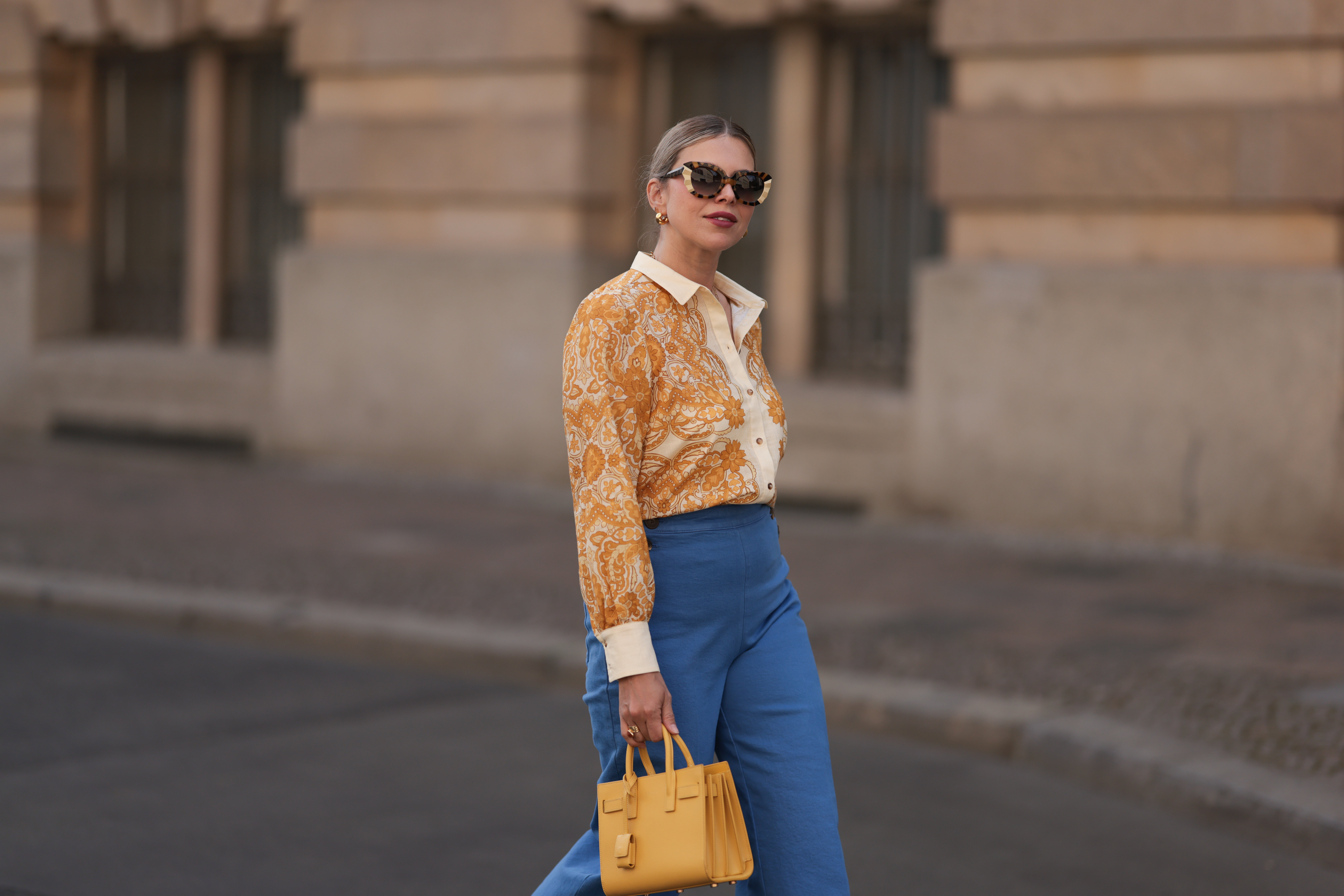 Aline Kaplan is seen wearing large sunglasses with beige, brown and black pattern from Tibi; chunky golden hoop earrings; several golden necklaces; a creme-white blouse with floral orange pattern from Sézane; a blue high-waisted pair of trousers with two button borders on the hips from Sézane; golden rings and a yellow leather handbag from Saint Laurent on June 07, 2024 in Berlin, Germany.