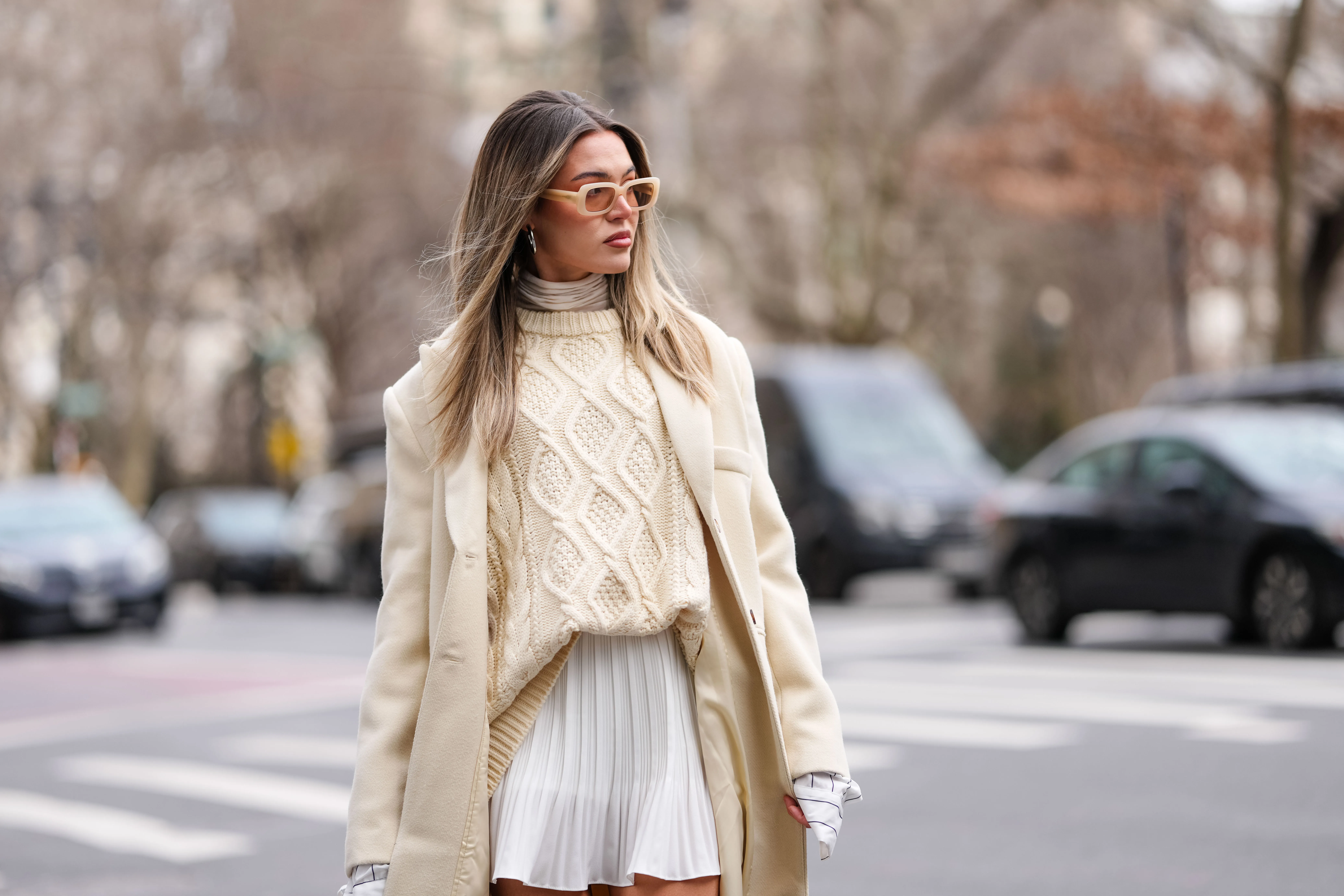 A guest wears yellow sunglasses, cream turtleneck long sleeve shirt, white pattern oversized buttoned up long sleeve shirt, cream knit oversized sweater, cream oversized wool coat, white mini skirt, outside Altuzarra, during the New York Fashion week Fall/Winter 2025 on February 8, 2025 in New York, New York.