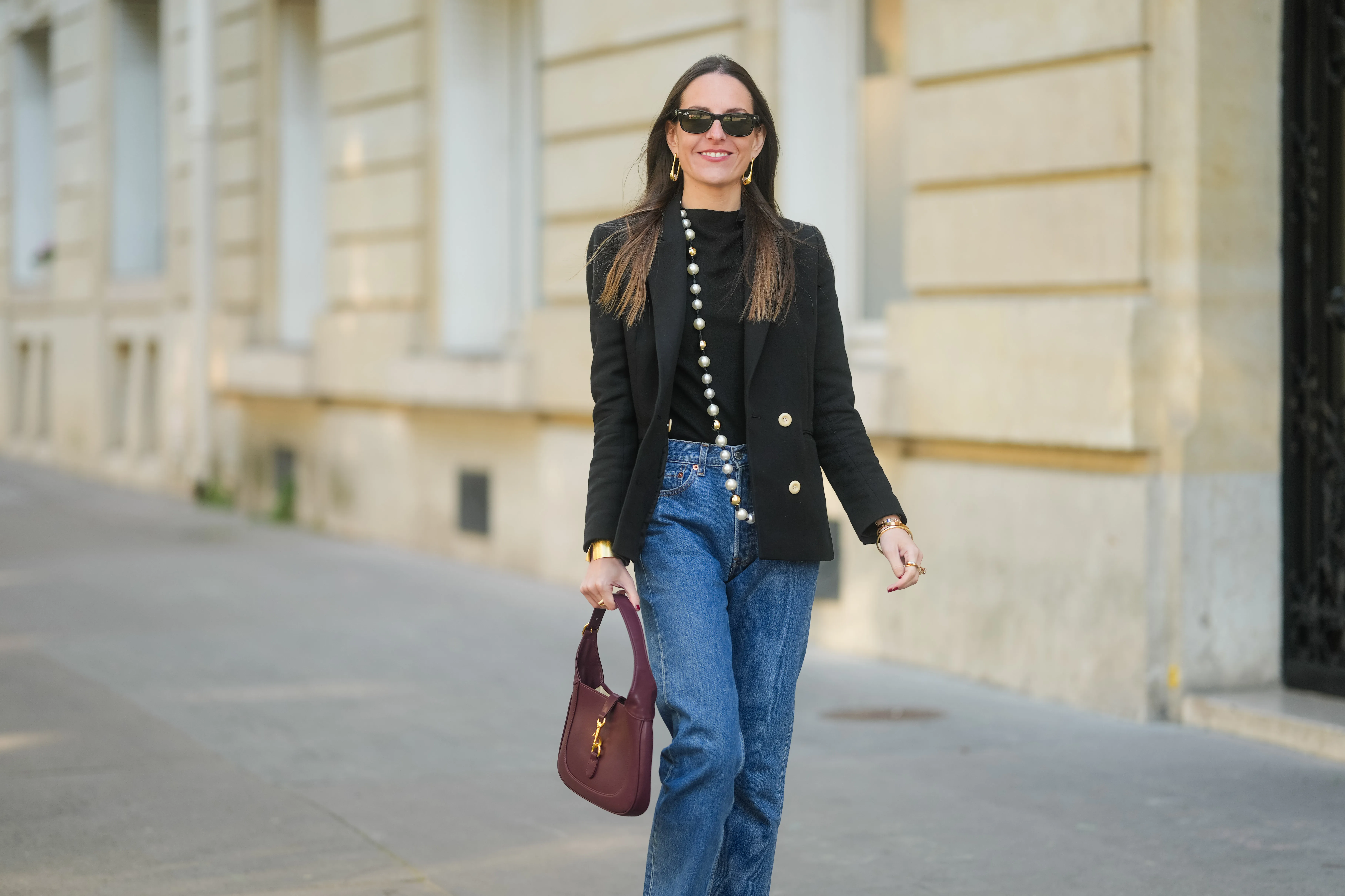 Alba Garavito Torre wears long straight brown hair styled with a center part. Black rectangular sunglasses are styled with large gold geometric earrings. A black T-shirt is layered under a tailored black double-breasted blazer with gold buttons by Sandro. A long strand of oversized pearls worn as a necklace by Sempiterno Paris is draped down the front. Classic blue Levi&rsquo;s 5015 straight-leg jeans are worn with the hem grazing the top of the shoe. A burgundy leather Jackie bag by Gucci is carried. Full body shot, during a street style fashion photo session, on April 09, 2025 in Paris, France.