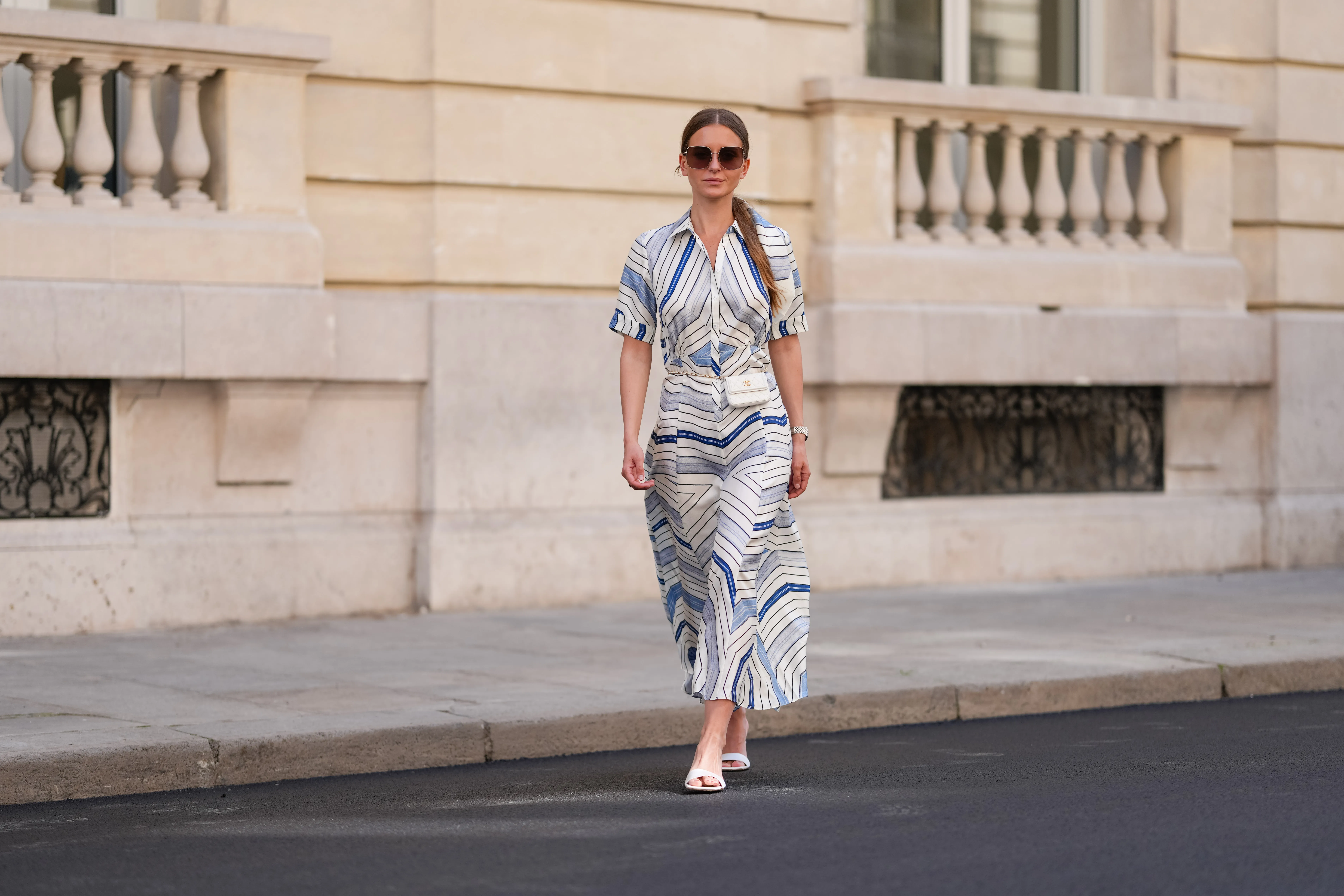 Diane Batoukina wears a white silk midi dress with blue and navy graphic stripe print, short sleeves, and a pointed collar by Sandro. A matching Sandro silk scarf is tied loosely around the neckline. A white mini flap bag with a chain strap and gold hardware by Chanel is worn crossbody. Footwear consists of white open-toe heeled sandals by Aeyde. Dior square sunglasses with dark gradient lenses complete the look. Long, straight brown hair is styled with a center part. Lips are subtly tinted and skin appears smooth and even. Full body shot, during a street style fashion photo session, on April 28, 2025 in Paris, France.