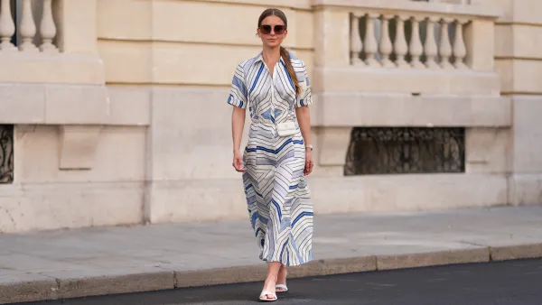 Diane Batoukina wears a white silk midi dress with blue and navy graphic stripe print, short sleeves, and a pointed collar by Sandro. A matching Sandro silk scarf is tied loosely around the neckline. A white mini flap bag with a chain strap and gold hardware by Chanel is worn crossbody. Footwear consists of white open-toe heeled sandals by Aeyde. Dior square sunglasses with dark gradient lenses complete the look. Long, straight brown hair is styled with a center part. Lips are subtly tinted and skin appears smooth and even. Full body shot, during a street style fashion photo session, on April 28, 2025 in Paris, France.