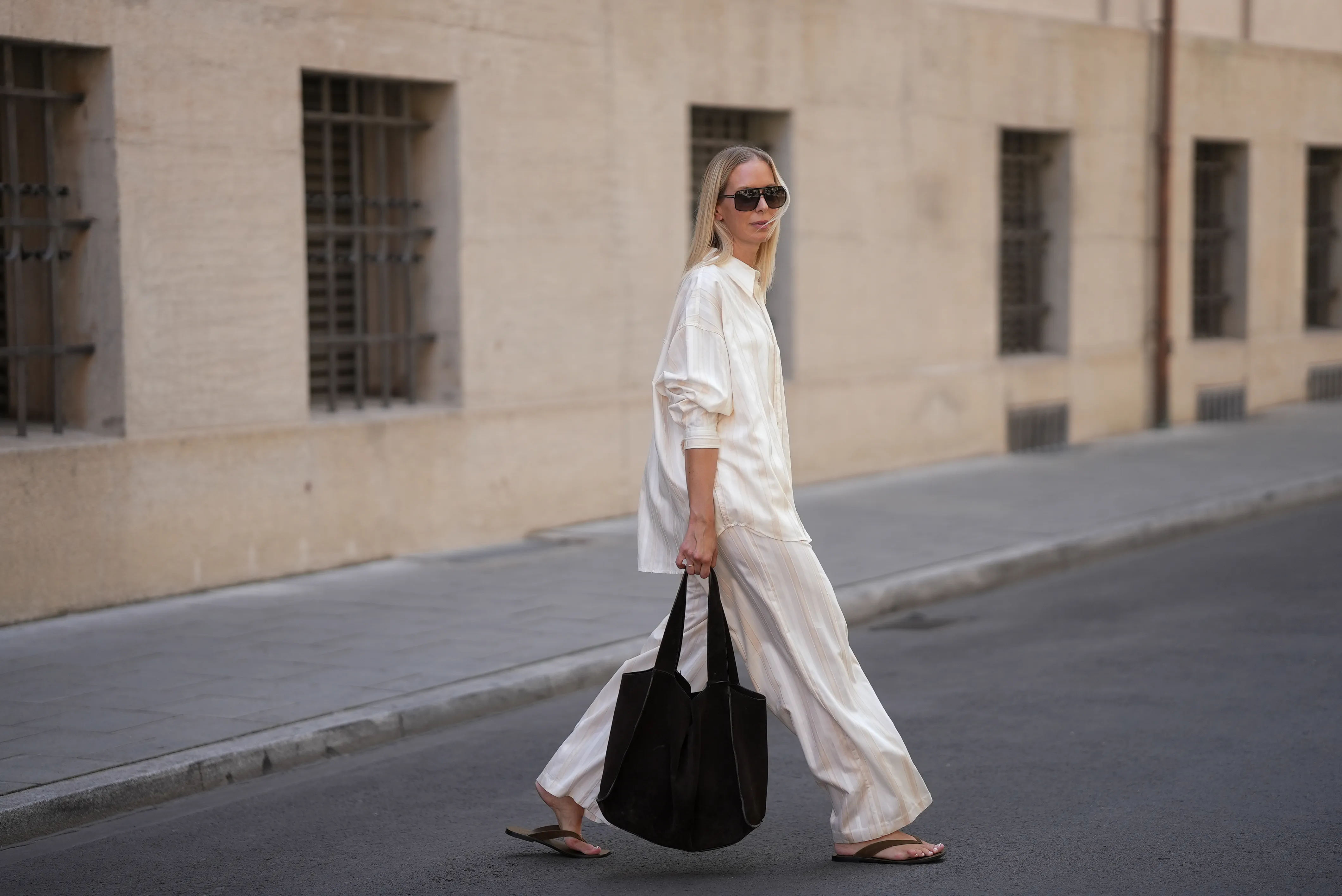 Mirja Klein is seen wearing a matching set consisting of a white striped oversized dobby-weave shirt with collar and buttons down in the front from H&M and a white striped straight pull-on trousers with loose fit and straight legs from H&M; a black suede tote bag with slouchy design and open top from COS; black oversized aviator sunglasses from Tom Ford; a pair of khaki green suede flip flops from A.EMERY on June 18, 2025 in Munich, Germany.