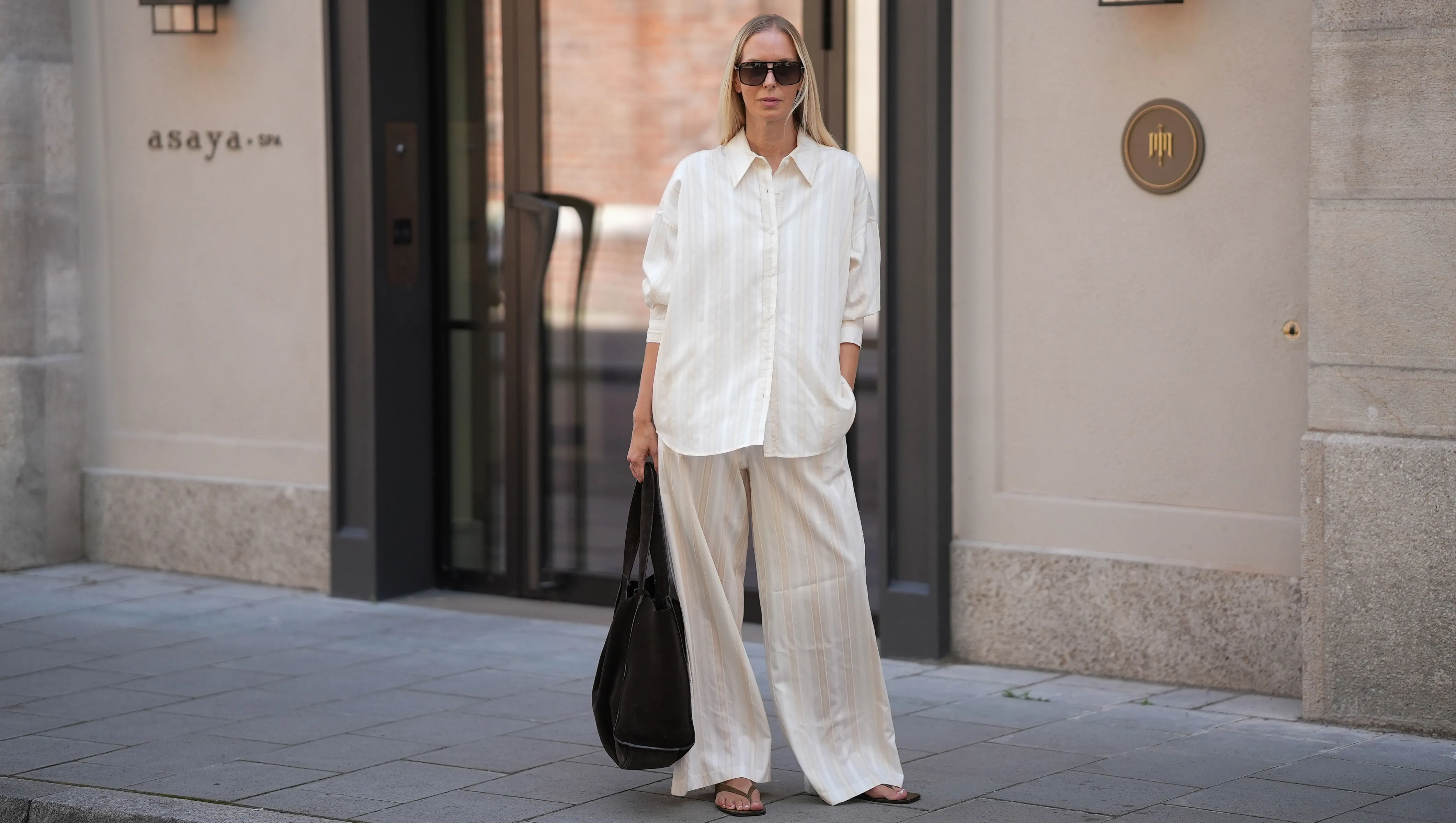 MUNICH, GERMANY - JUNE 18: Mirja Klein is seen wearing a matching set consisting of a white striped oversized dobby-weave shirt with collar and buttons down in the front from H&M and a white striped straight pull-on trousers with loose fit and straight legs from H&M; a black suede tote bag with slouchy design and open top from COS; black oversized aviator sunglasses from Tom Ford; a pair of khaki green suede flip flops from A.EMERY on June 18, 2025 in Munich, Germany. (Photo by Moritz Scholz/Getty Images)