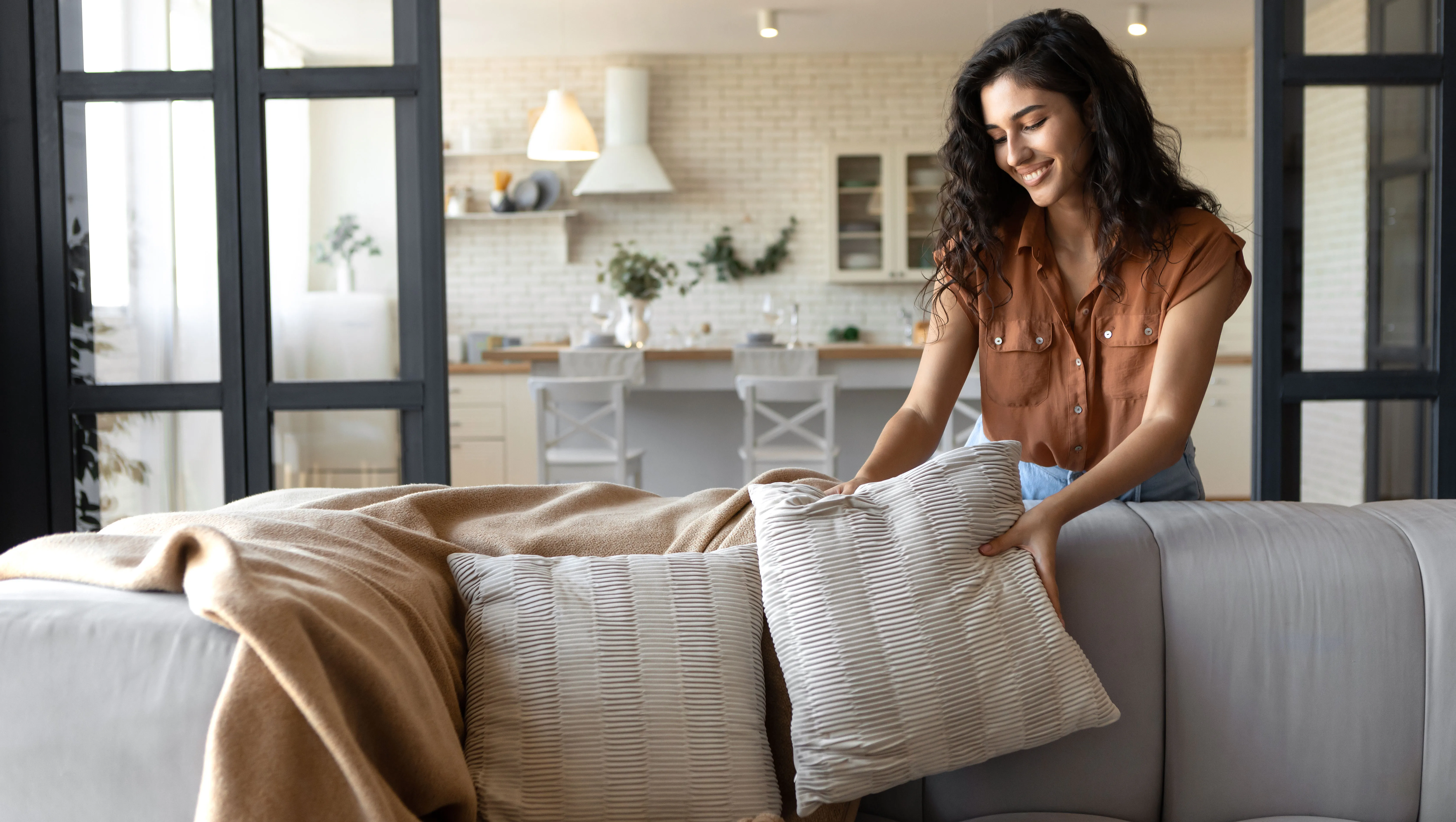 Lovely young woman putting soft pillows and plaid on comfy sofa, making her home cozy and warm, copy space. Millennial lady decorating her couch with cushions and blanket for autumn or winter