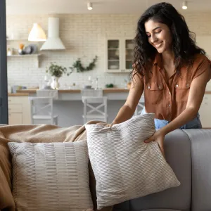 Lovely young woman putting soft pillows and plaid on comfy sofa, making her home cozy and warm, copy space. Millennial lady decorating her couch with cushions and blanket for autumn or winter