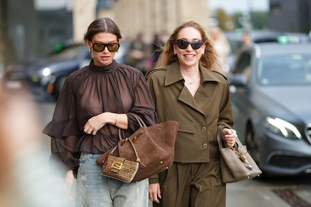 COPENHAGEN, DENMARK - AUGUST 06: A guest wears a sheer, dark brown blouse with a high neckline and layered ruffle sleeves. The hair is styled in a sleek, center-parted updo. Sunglasses are oversized with a rectangular frame. Earrings are small and circular. A bracelet is worn on the wrist. The jeans are light blue with a distressed finish. A large brown suede bag is carried, featuring gold hardware. A smaller brown Fendi monogrammed bag with a top handle is also visible. Another guest wears sunglasses with dark lenses and a brown frame. The hair is styled in loose waves. A brown double-breasted jacket features a notched lapel, button closure, and a flap pocket on one side. The sleeves are rolled up. Matching high-waisted trousers have a pleated front and a relaxed fit. A taupe textured handbag is carried, with short handles, outside Gestuz, during Copenhagen Fashion Week, on August 06, 2025 in Copenhagen, Denmark (Photo by Edward Berthelot/Getty Images)